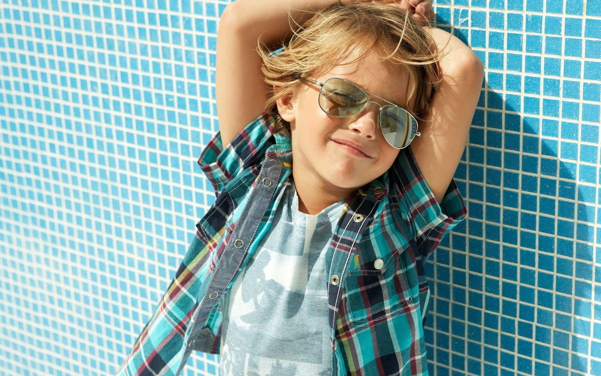 Cute Boy 桌布 壁紙 — 一個微笑的孩子戴著太陽眼鏡，在陽光明媚的日子裡靠著藍色磁磚泳池牆。