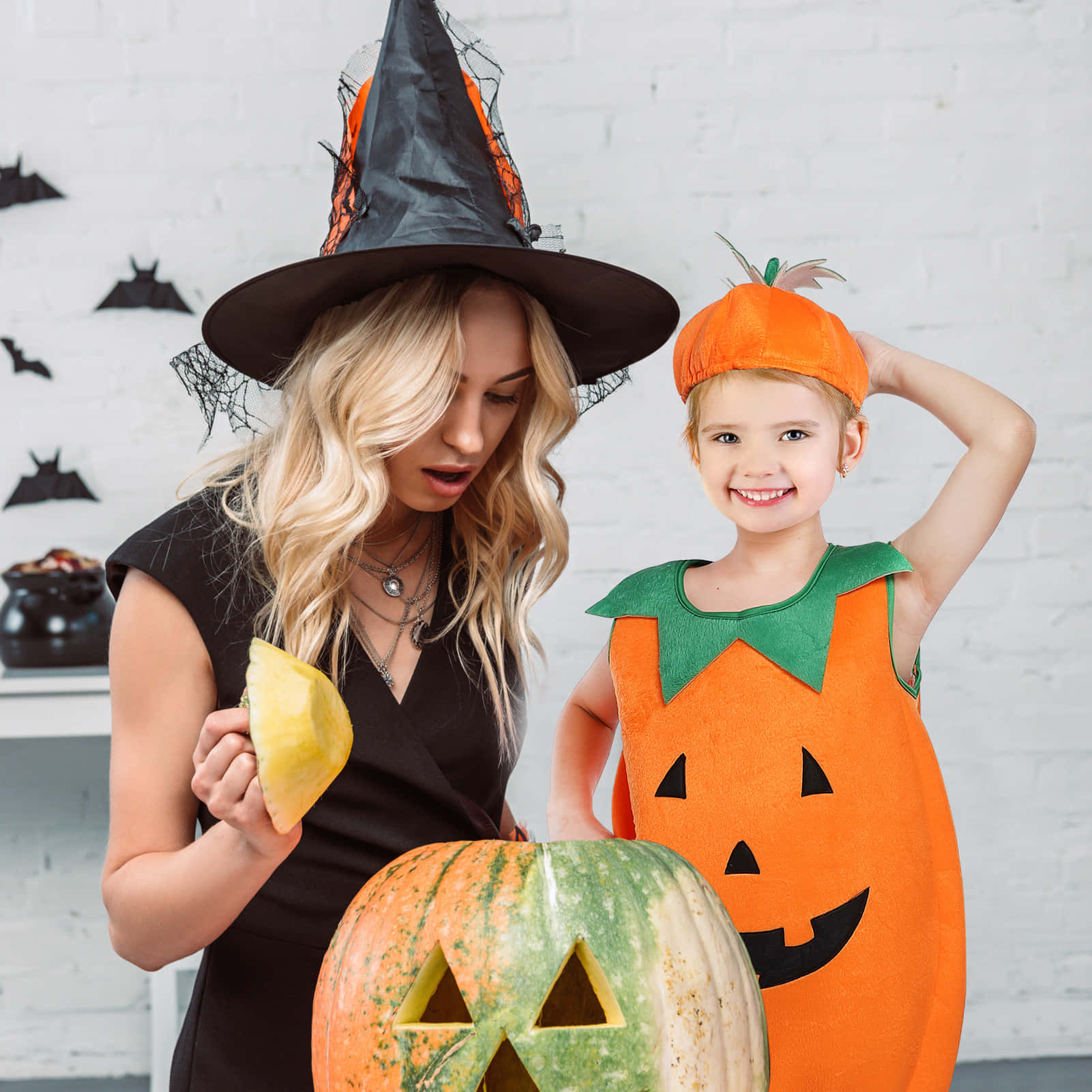 Halloween Costume 桌布 壁紙 — 一個微笑的孩子穿著南瓜服裝，站在一個戴著巫婆帽的女人旁邊，正在雕刻一個大南瓜燈。