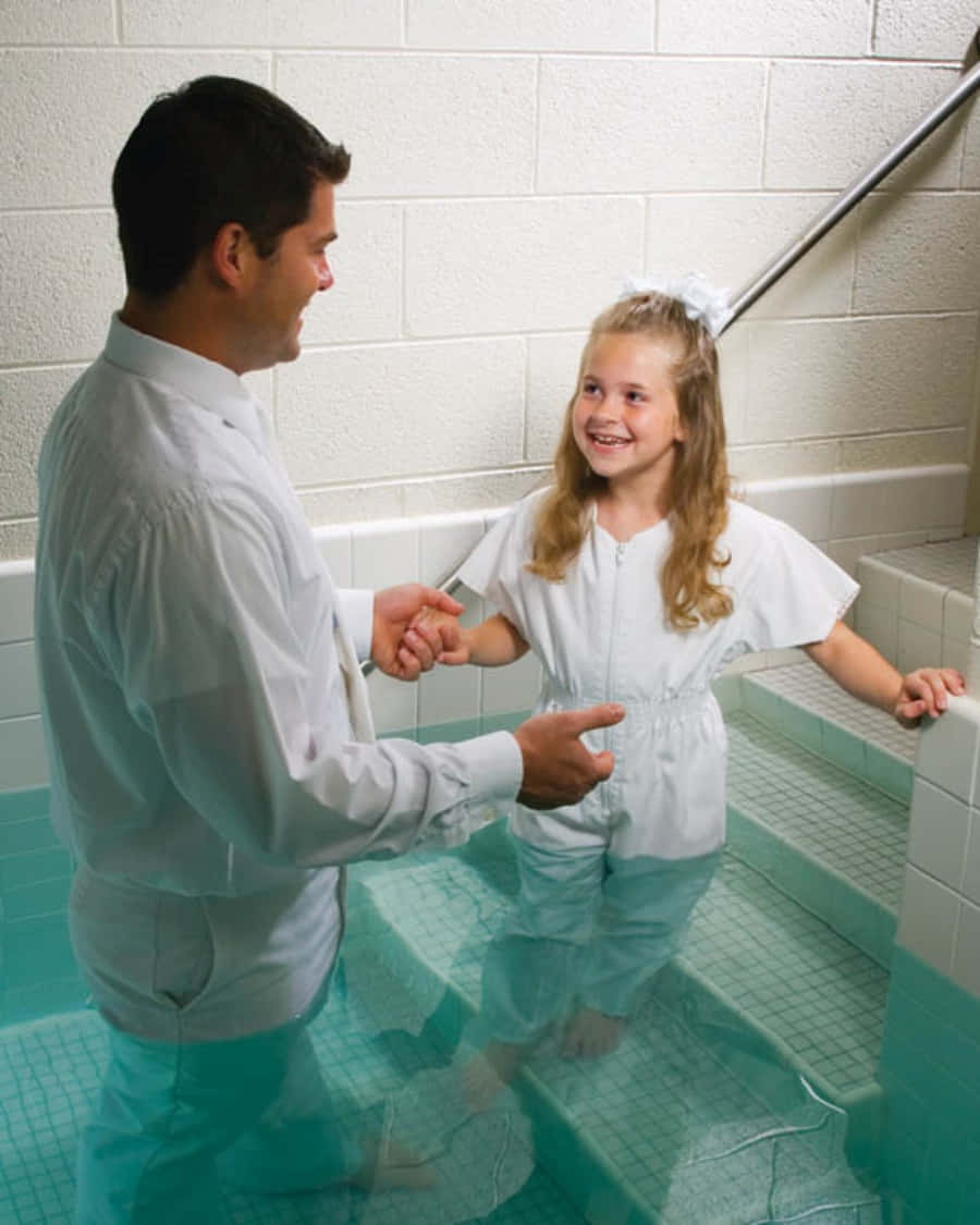 Baptism 桌布 壁紙 — 一個微笑的小女孩穿著白色洗禮袍，在男人的引導下踏入瓷磚洗禮池。