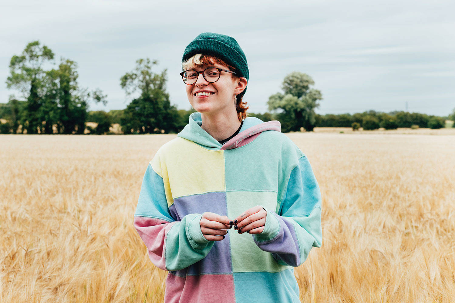 Cavetown 桌布 壁紙 — 一個微笑的年輕人穿著彩色拼布連帽衫和綠色無邊帽，站在陰天下的金色麥田裡。