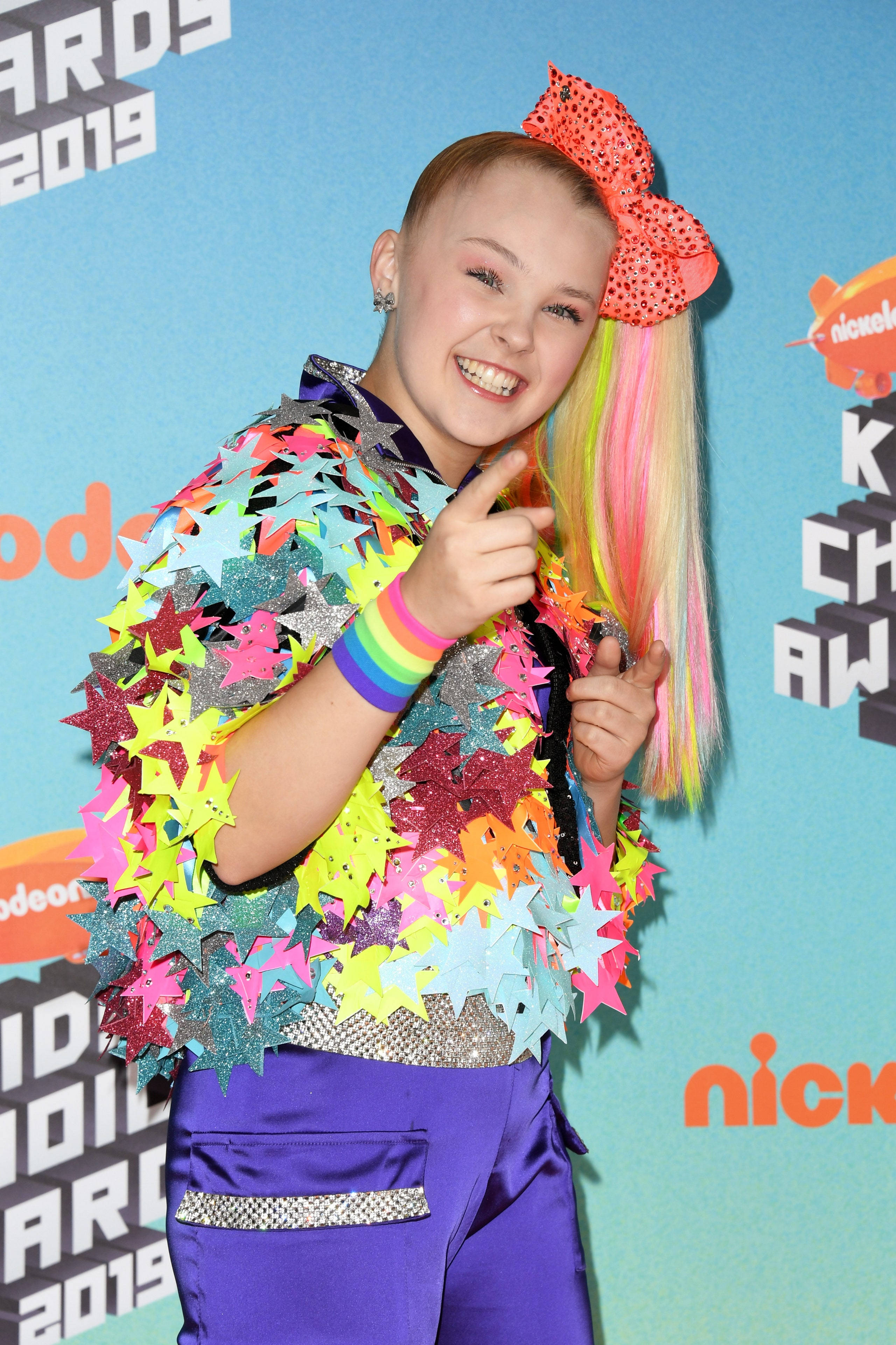 JoJo Siwa 桌布 壁紙 — 一個微笑的年輕女孩，彩虹頭髮和一個巨大的蝴蝶結，穿著閃閃發光的星星外套出席2019年兒童選擇獎。