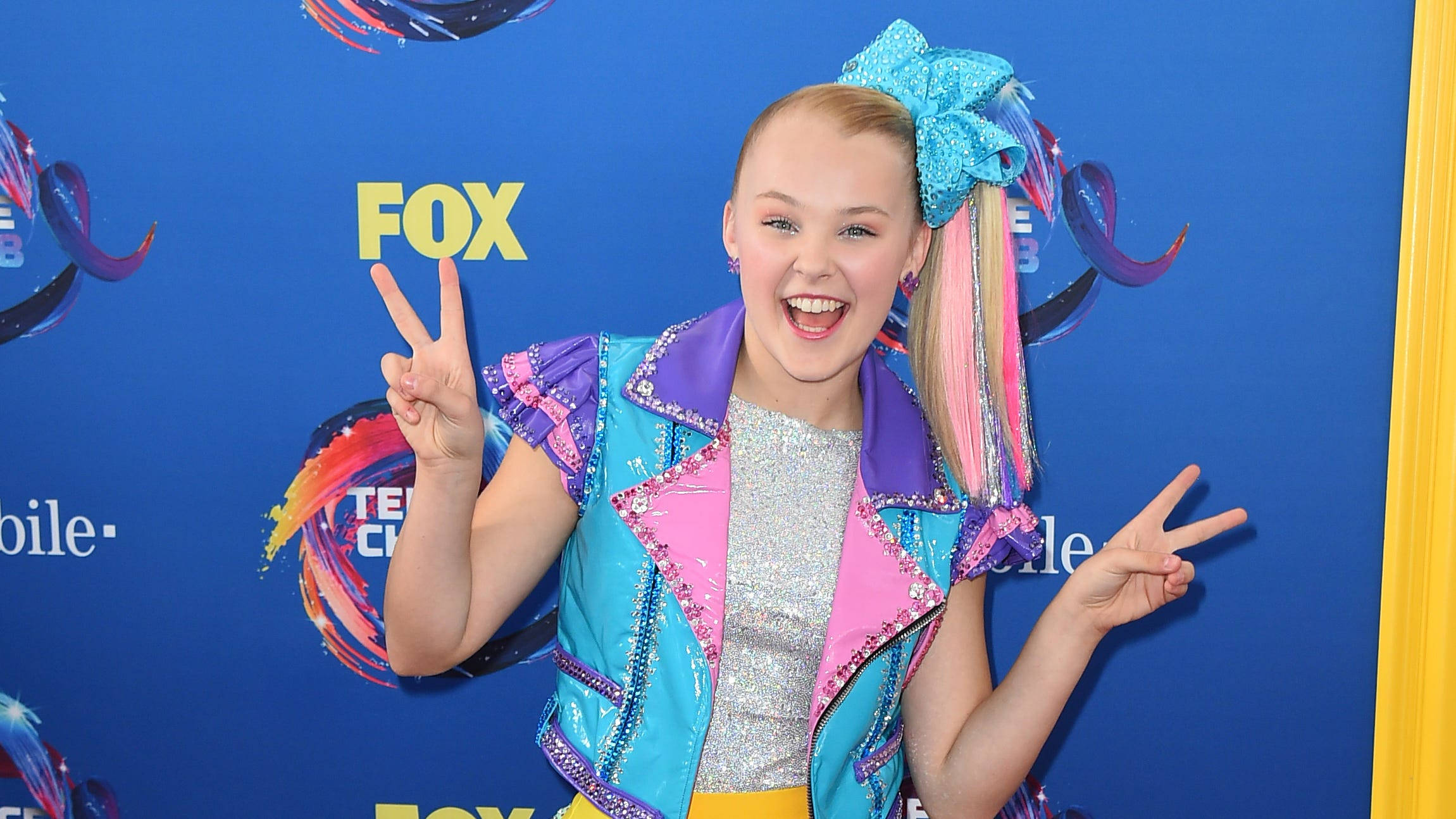 JoJo Siwa 桌布 壁紙 — 一個微笑的年輕女孩，戴著一個大藍色蝴蝶結和色彩繽紛的頭髮，在藍色FOX活動背景前做出和平手勢。