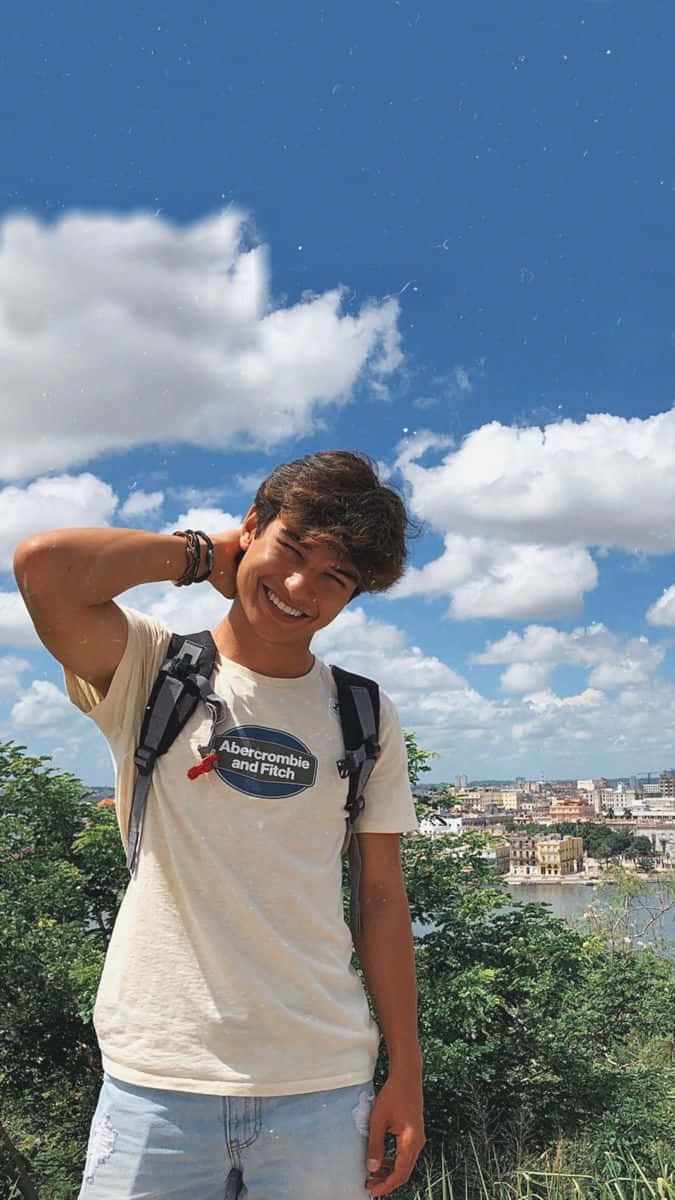 Boy 桌布 壁紙 — 一個微笑的年輕男子揹著背包，站在可以俯瞰沿海城市的小山上，天空湛藍，白雲朵朵。