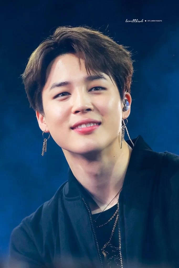 BTS 智旻 桌布 壁紙 — 一個微笑的年輕男子特寫肖像，擁有造型頭髮和垂墜耳環，背景是深藍色舞台。