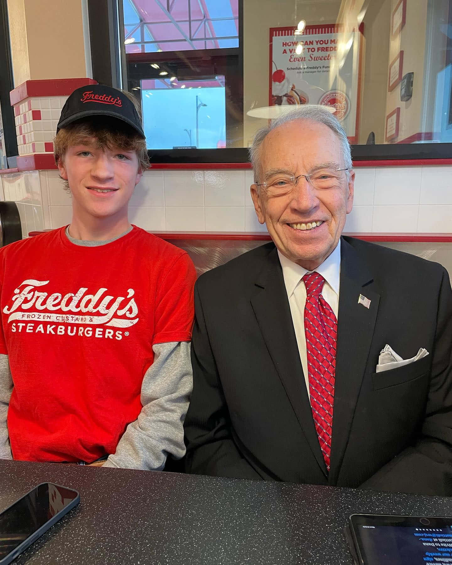政治 桌布 壁紙 — 一個微笑的年輕男子穿著Freddy's襯衫，與一位穿著西裝的年長紳士坐在餐廳卡座旁。