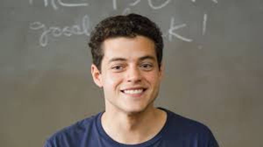 Rami Malek 桌布 壁紙 — 一個微笑的年輕男子穿著藍色襯衫，站在一塊用白色粉筆寫著「好運！」的黑板前。