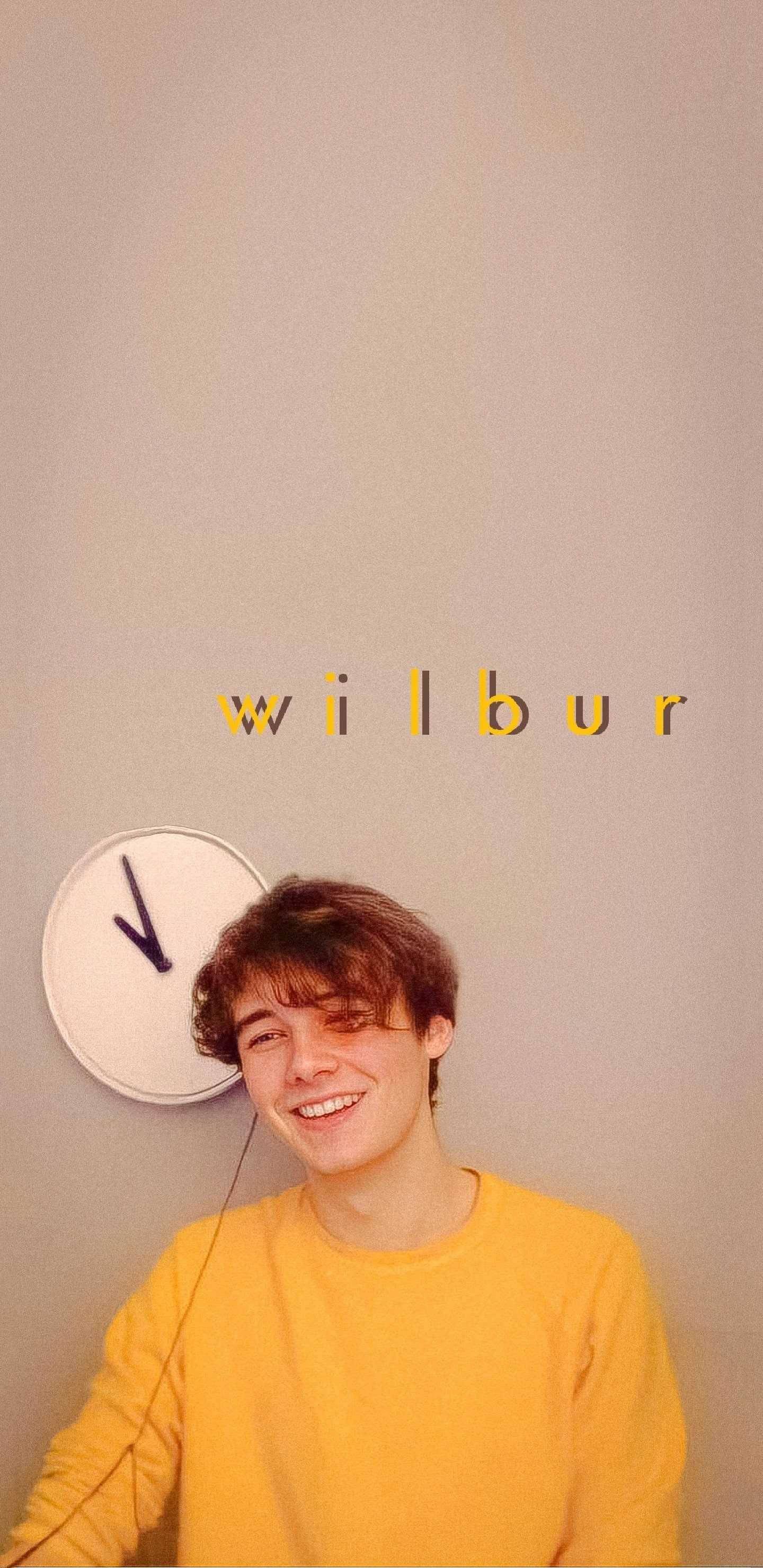 Wilbur 桌布 壁紙 — 一個微笑的年輕男子穿著黃色毛衣，站在掛牆時鐘和黃色字母wilbur前面。