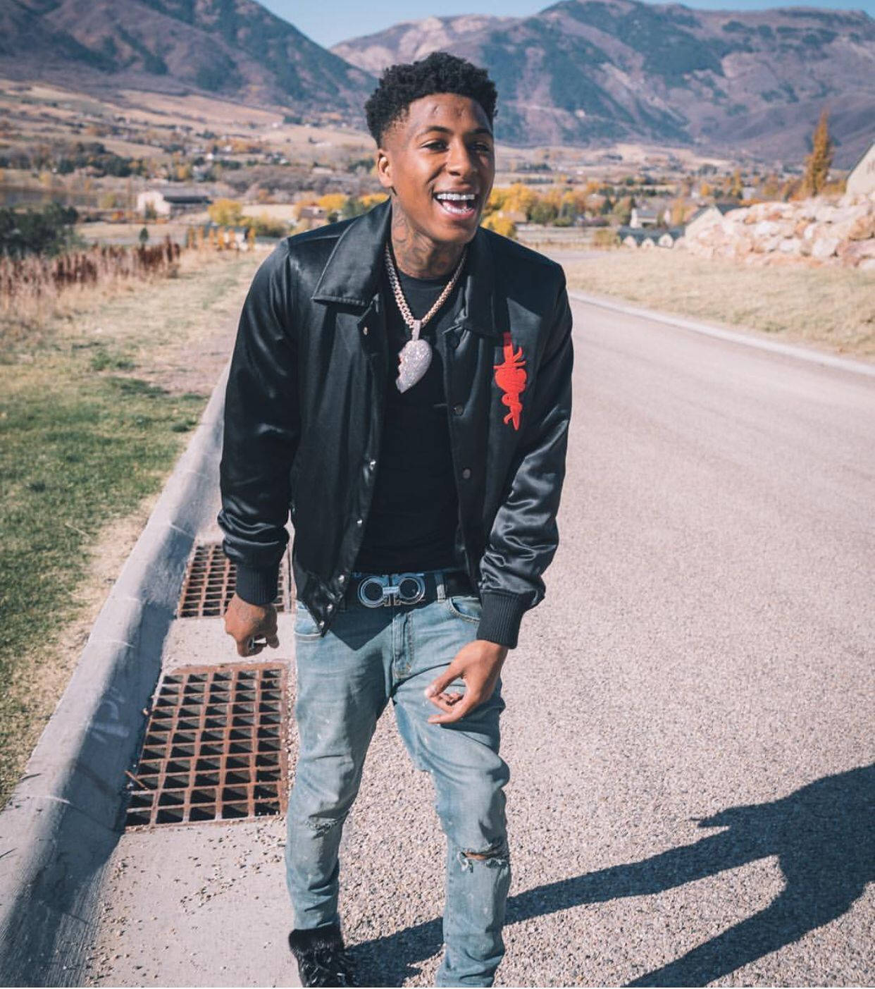 Youngboy 桌布 壁紙 — 一個微笑的年輕男子穿著黑色皮夾克和破洞牛仔褲，站在路邊，背景是山脈和秋天的樹木。