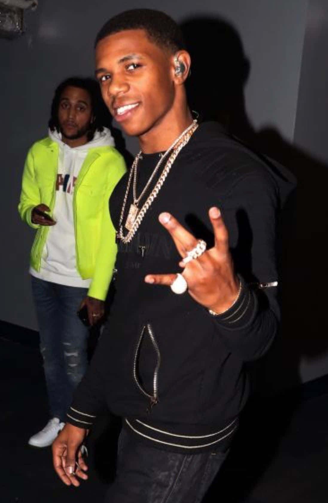A Boogie Wit Da Hoodie 桌布 壁紙 — 一個微笑的年輕男子穿著黑色連帽衫和金色鏈條，比出和平手勢，而另一個穿著霓虹外套的男子站在他身後，在一個燈光昏暗的房間裡。
