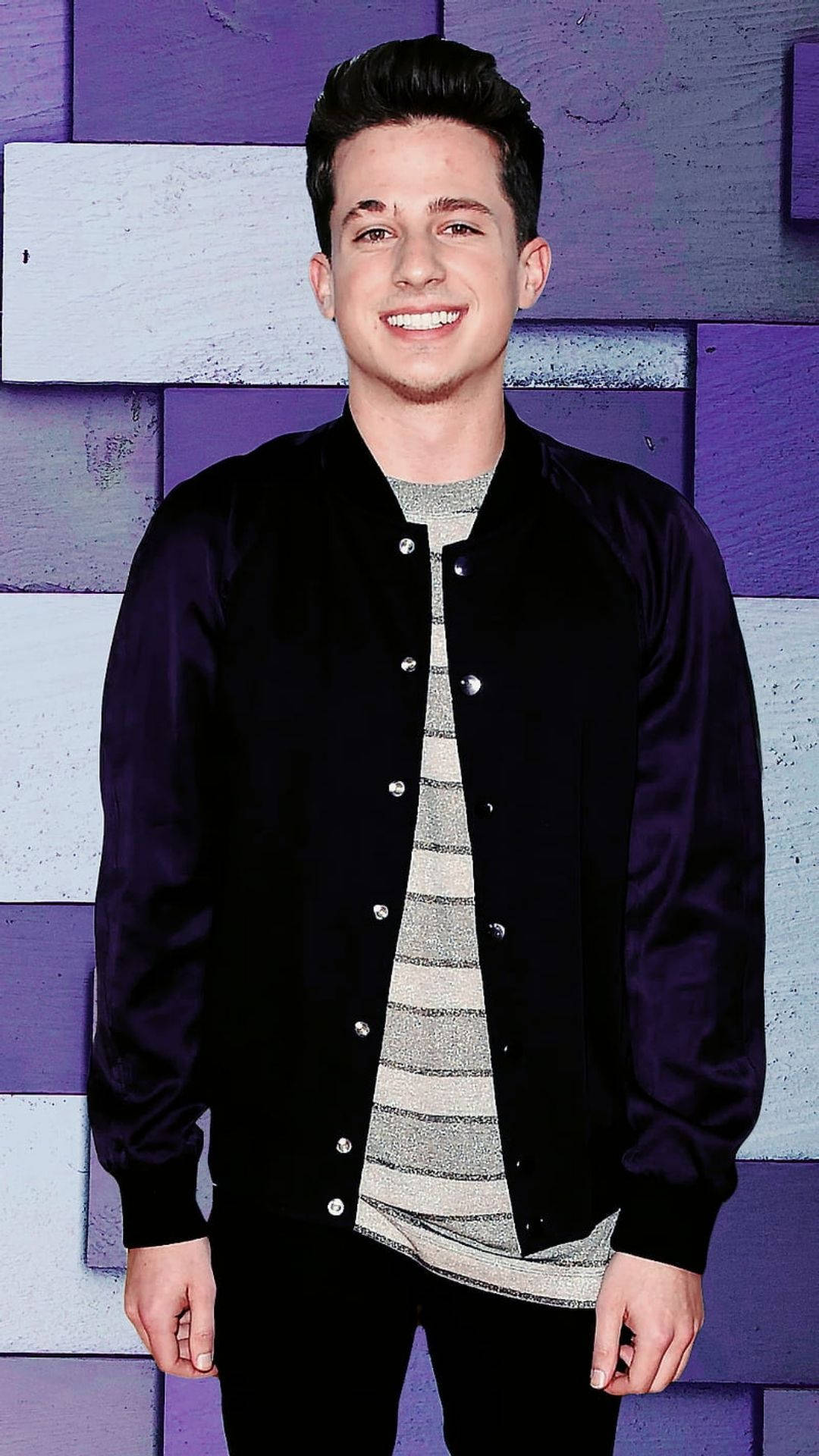 Charlie Puth 桌布 壁紙 — 一個微笑的年輕男子穿著黑色飛行員夾克，站在紫色和白色的木板牆前。