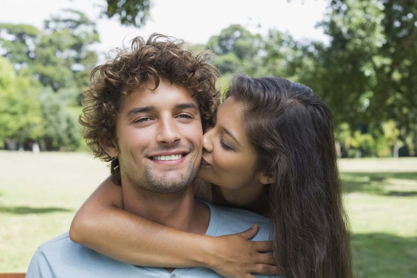 Boyfriend And Girlfriend 桌布 壁紙 — 一個微笑的捲髮男人在陽光明媚的公園裡被一個擁抱他的女人親吻臉頰。