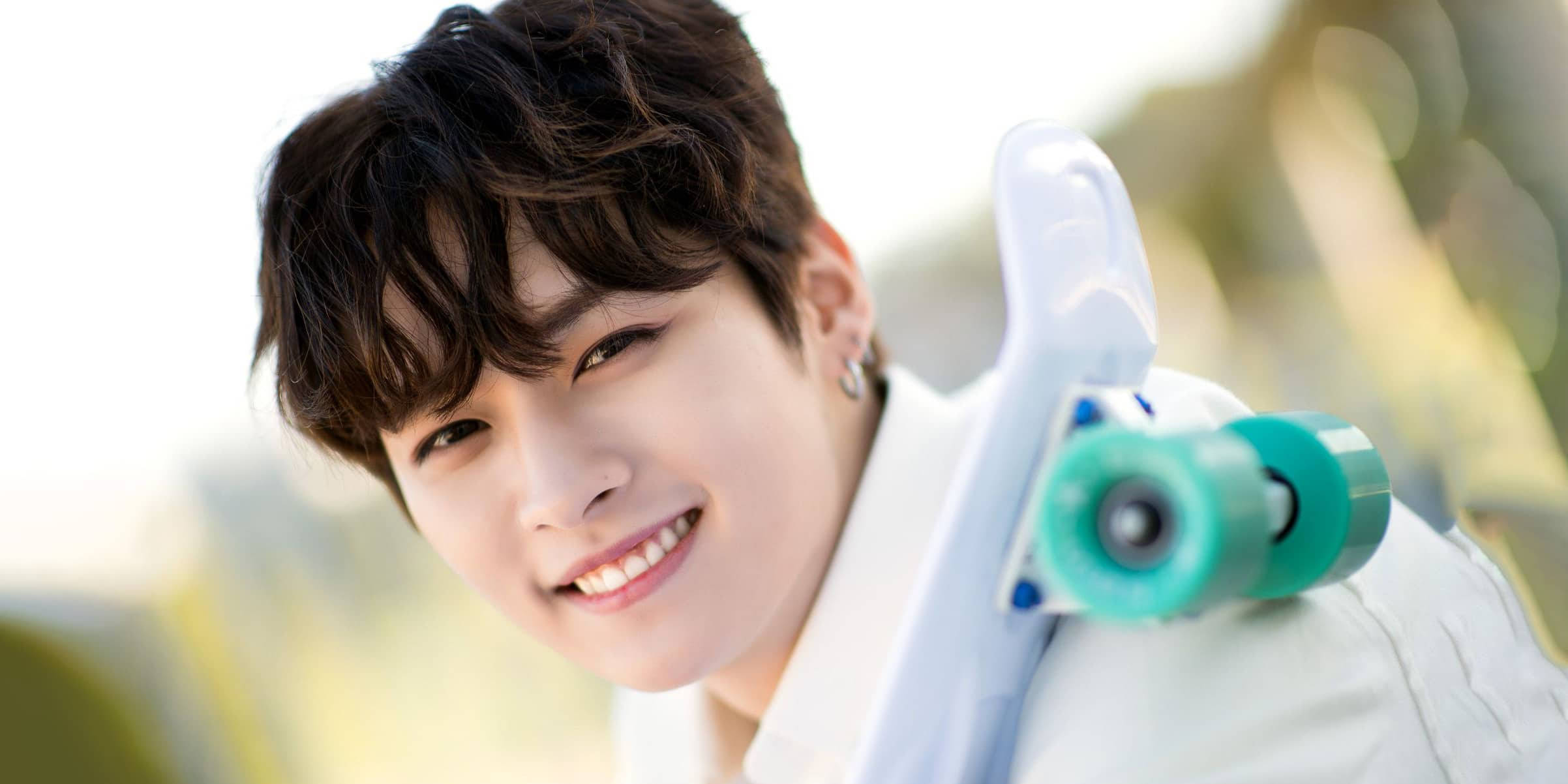 Stray Kids 桌布 壁紙 — 一個微笑的深色頭髮年輕男子，在明亮模糊的戶外背景下，將滑板扛在肩上。