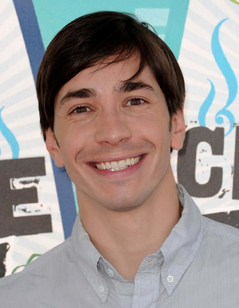Justin Long 桌布 壁紙 — 一個微笑的深色頭髮年輕男子的特寫肖像，穿著淺藍色鈕扣襯衫，背景是色彩繽紛的圖形背景。