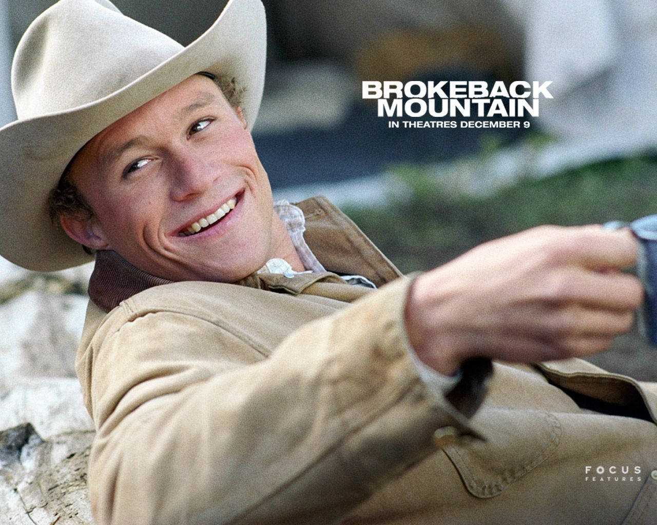 brokeback mountain 桌布 壁紙 — 一個微笑的牛仔，戴著棕褐色帽子和外套，靠著岩石向後傾，手持一個帶有電影標題疊加的杯子。