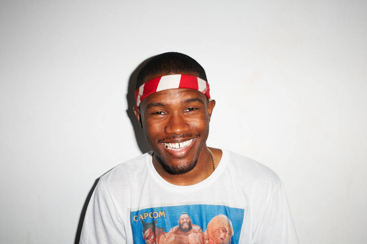 Frank Ocean 桌布 壁紙 — 一個微笑的男人，戴著紅白條紋頭帶，穿著印有Capcom電玩圖案的白色T恤，背景是純白牆壁。