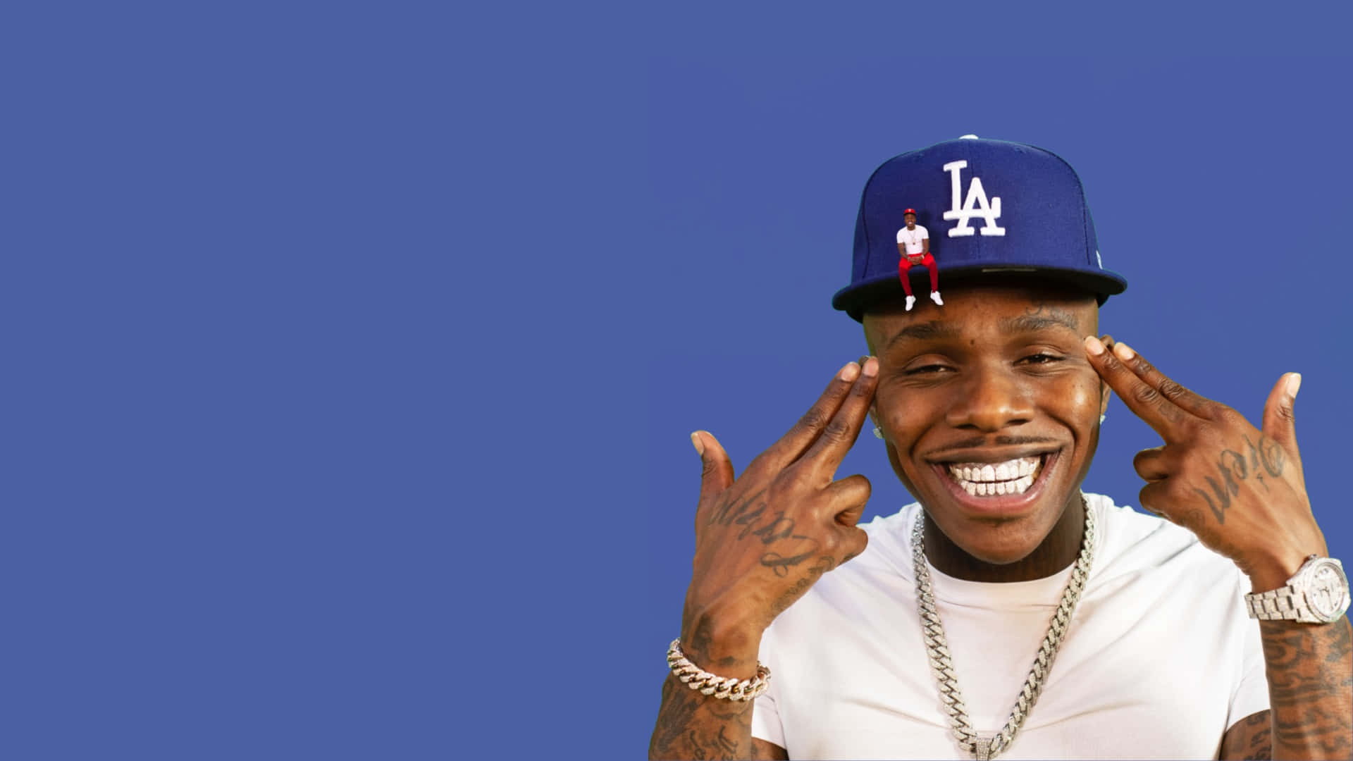Dababy 桌布 壁紙 — 一個微笑的男人，戴著藍色LA帽，上面有一個小公仔，雙手框著臉，背景是純藍色。