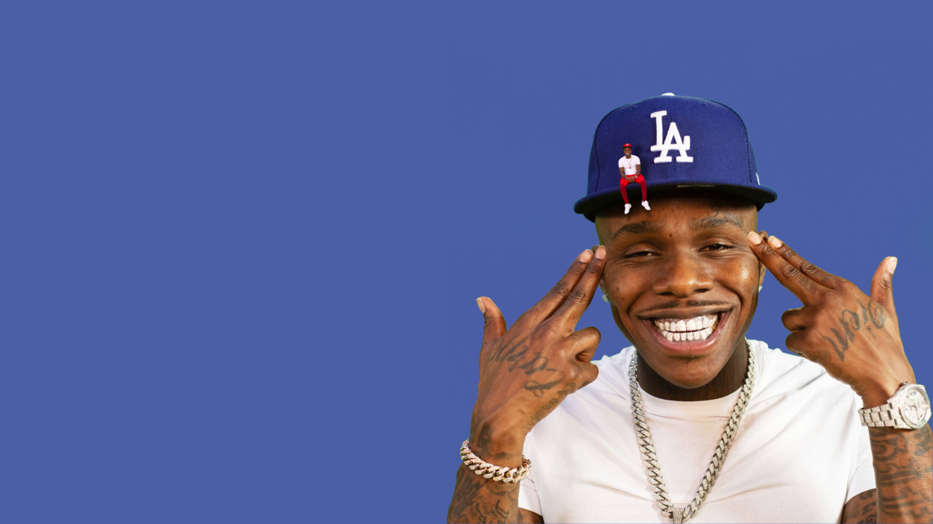 Dababy 桌布 壁紙 — 一個微笑的男人，戴著藍色LA帽子，上面有一個小公仔，雙手框著臉，背景是純藍色。