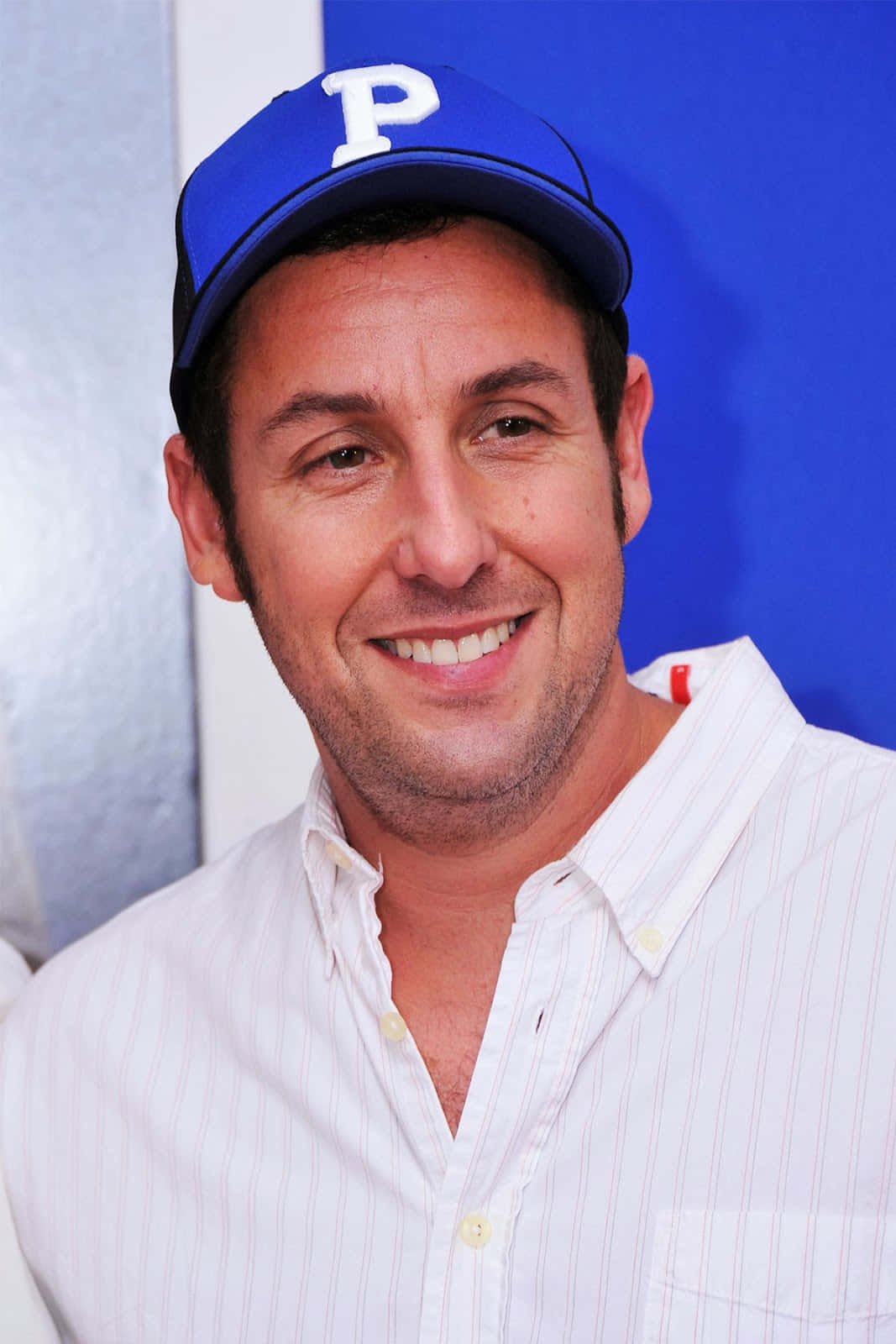 Adam Sandler 桌布 壁紙 — 一個微笑的男人，戴著藍色帽子，上面有白色「P」，身穿白色條紋襯衫，背景為藍色和銀色。