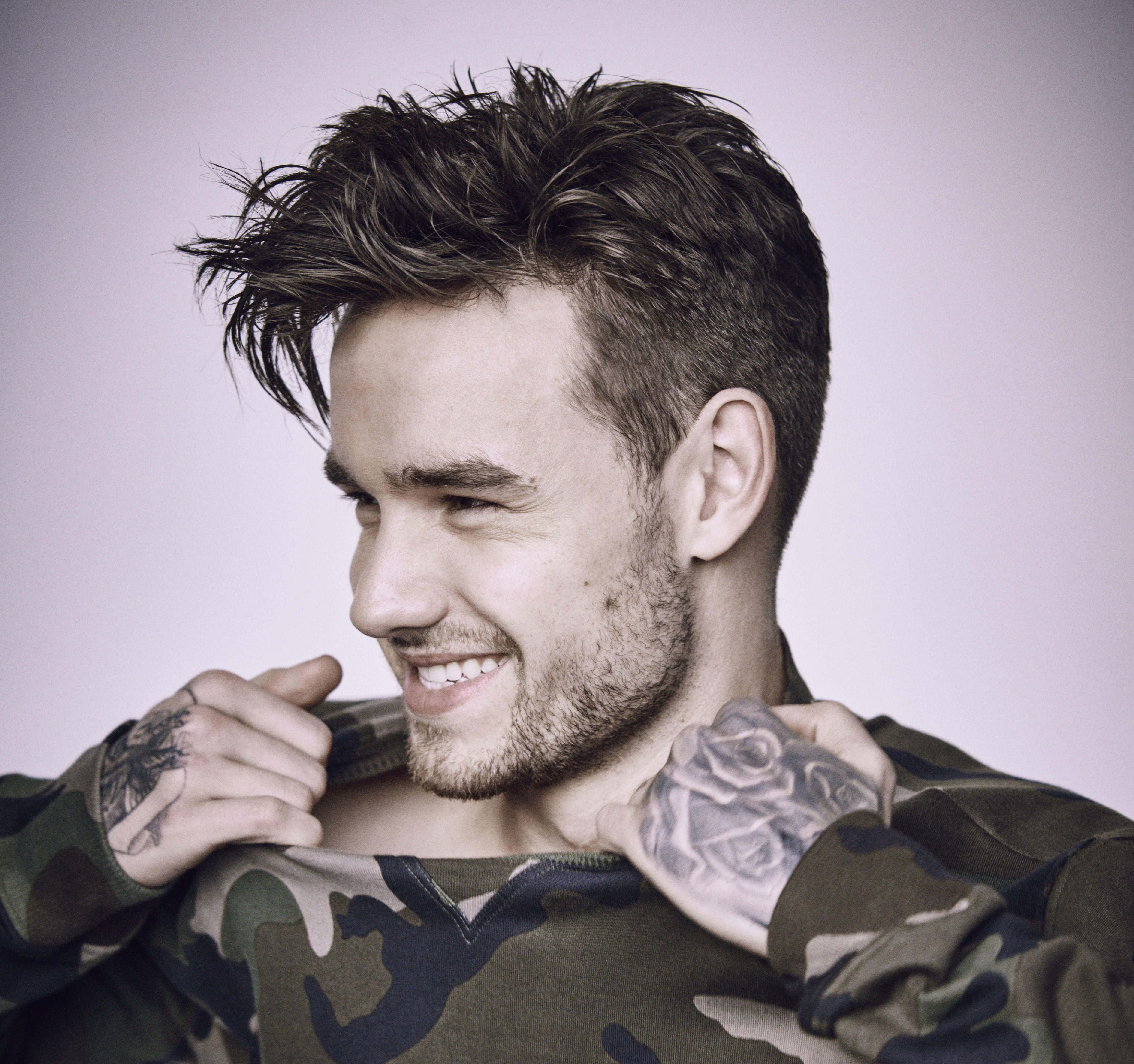 liam payne 桌布 壁紙 — 一個微笑的男人，手上有刺青，在柔和的紫色背景前掀起迷彩襯衫露出胸膛。