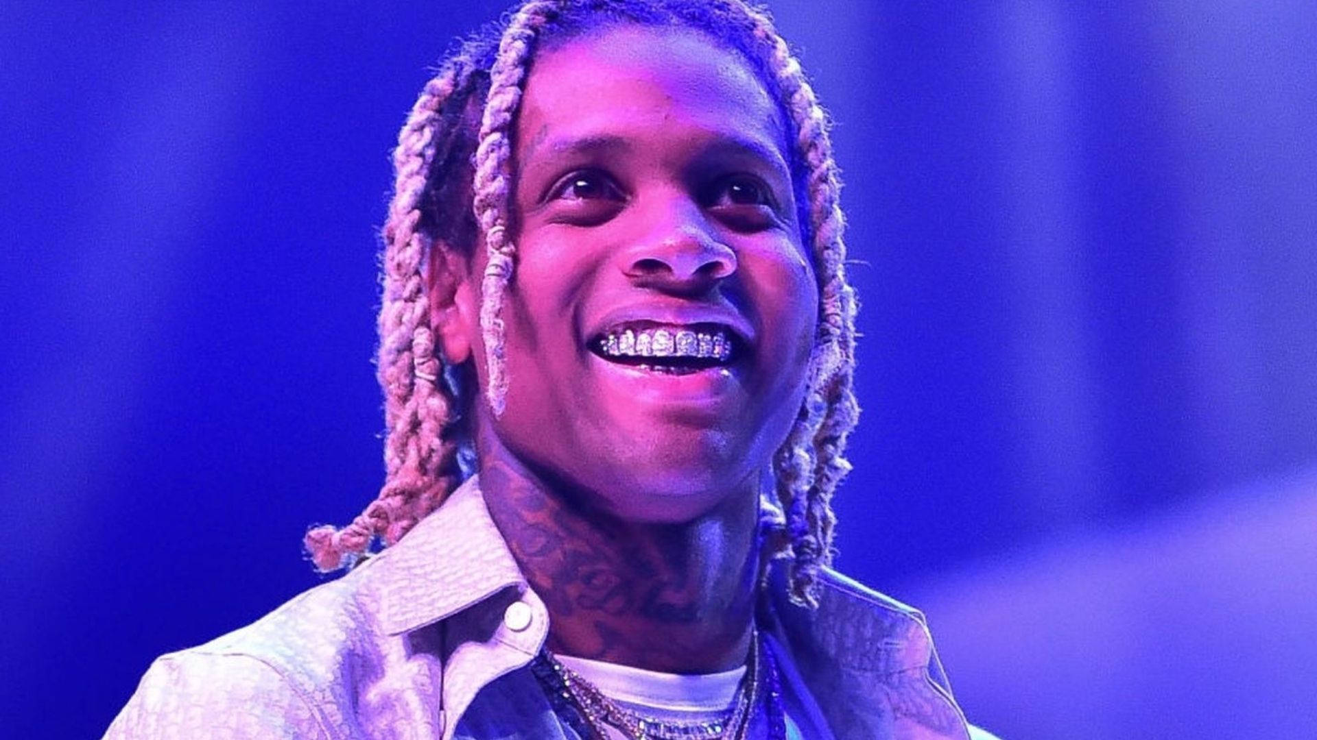 Lil Durk 桌布 壁紙 — 一個微笑的男人，梳著辮子頭，戴著鑽石牙套，在充滿活力的藍色舞台燈光下。