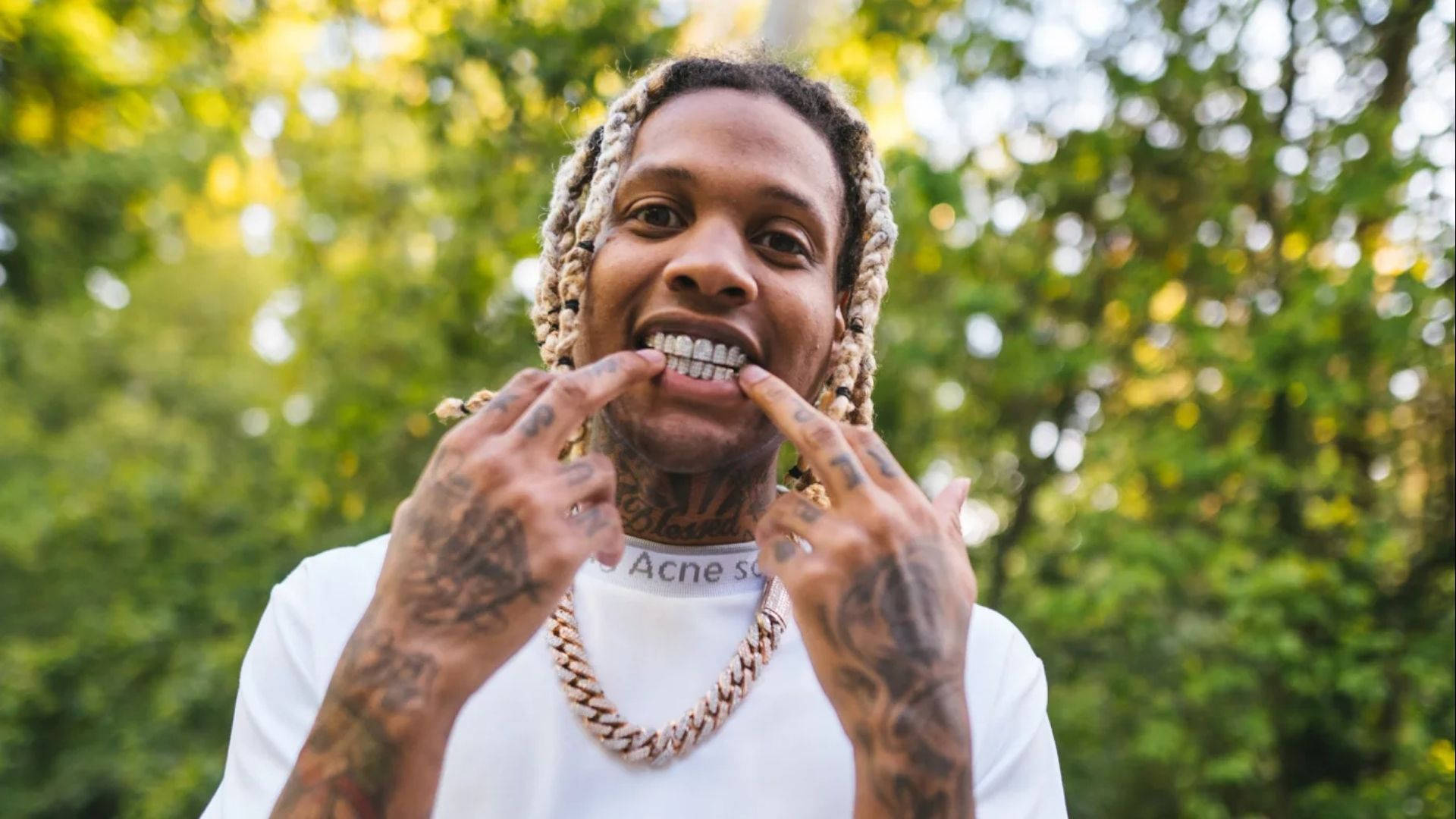 Lil Durk 桌布 壁紙 — 一個微笑的男人，梳著辮子頭和刺青，在陽光普照的綠色森林背景下，頑皮地指著他的金牙套。