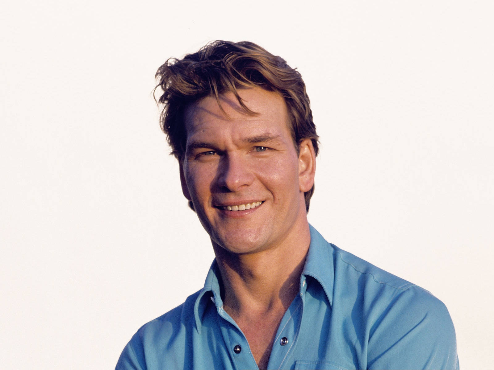 Patrick Swayze 桌布 壁紙 — 一個微笑的男人，棕色頭髮凌亂，穿著淺藍色鈕扣襯衫，背景是純白色。