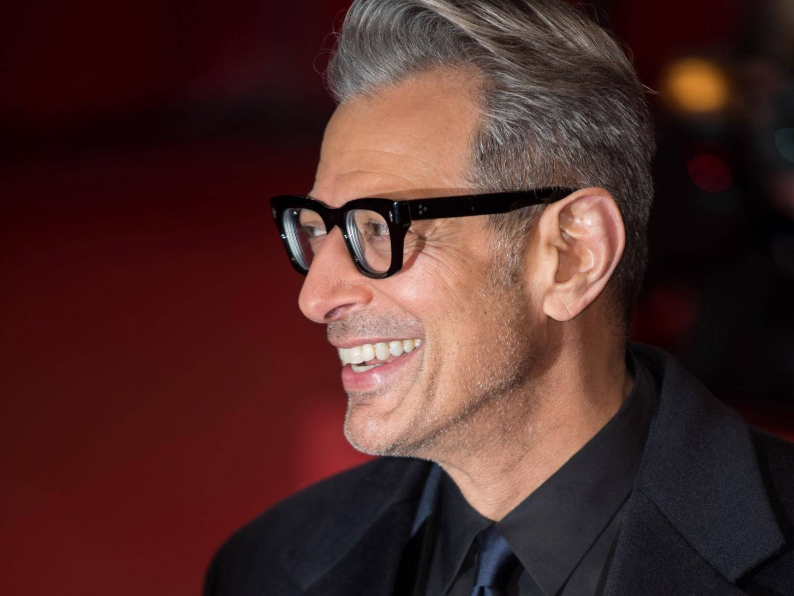 jeff goldblum 桌布 壁紙 — 一個微笑的男人，灰白頭髮，戴黑色眼鏡，背景是模糊的紅色。