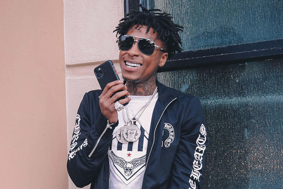 Youngboy 桌布 壁紙 — 一個微笑的男人，留著辮子頭，戴著飛行員太陽眼鏡，靠著牆壁，手裡拿著手機。
