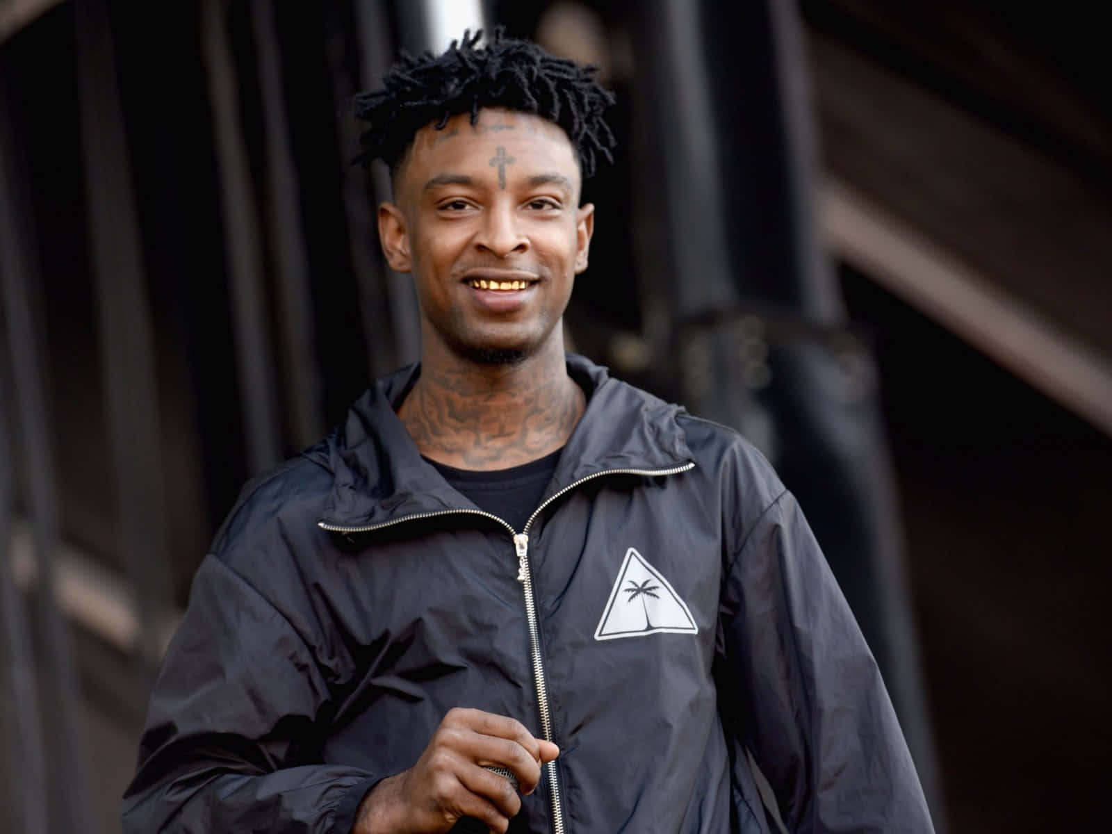 21 Savage 桌布 壁紙 — 一個微笑的男人，留著辮子頭和刺青，穿著黑色外套，上面有白色棕櫚樹標誌，背景是黑暗的城市。