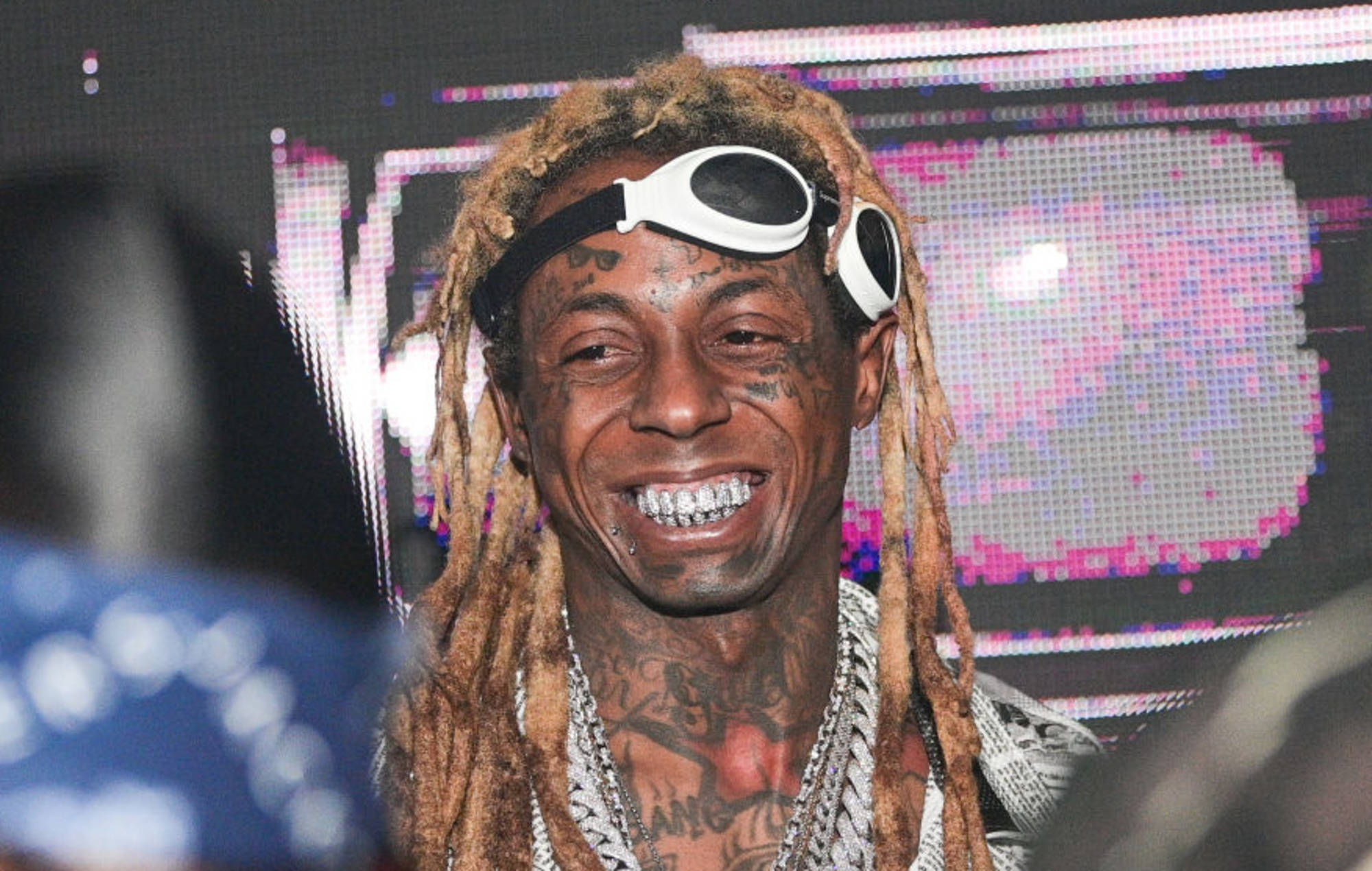 Lil Wayne 桌布 壁紙 — 一個微笑的男人，留著辮子頭和護目鏡，在發光的像素背景前對著鏡頭咧嘴笑。
