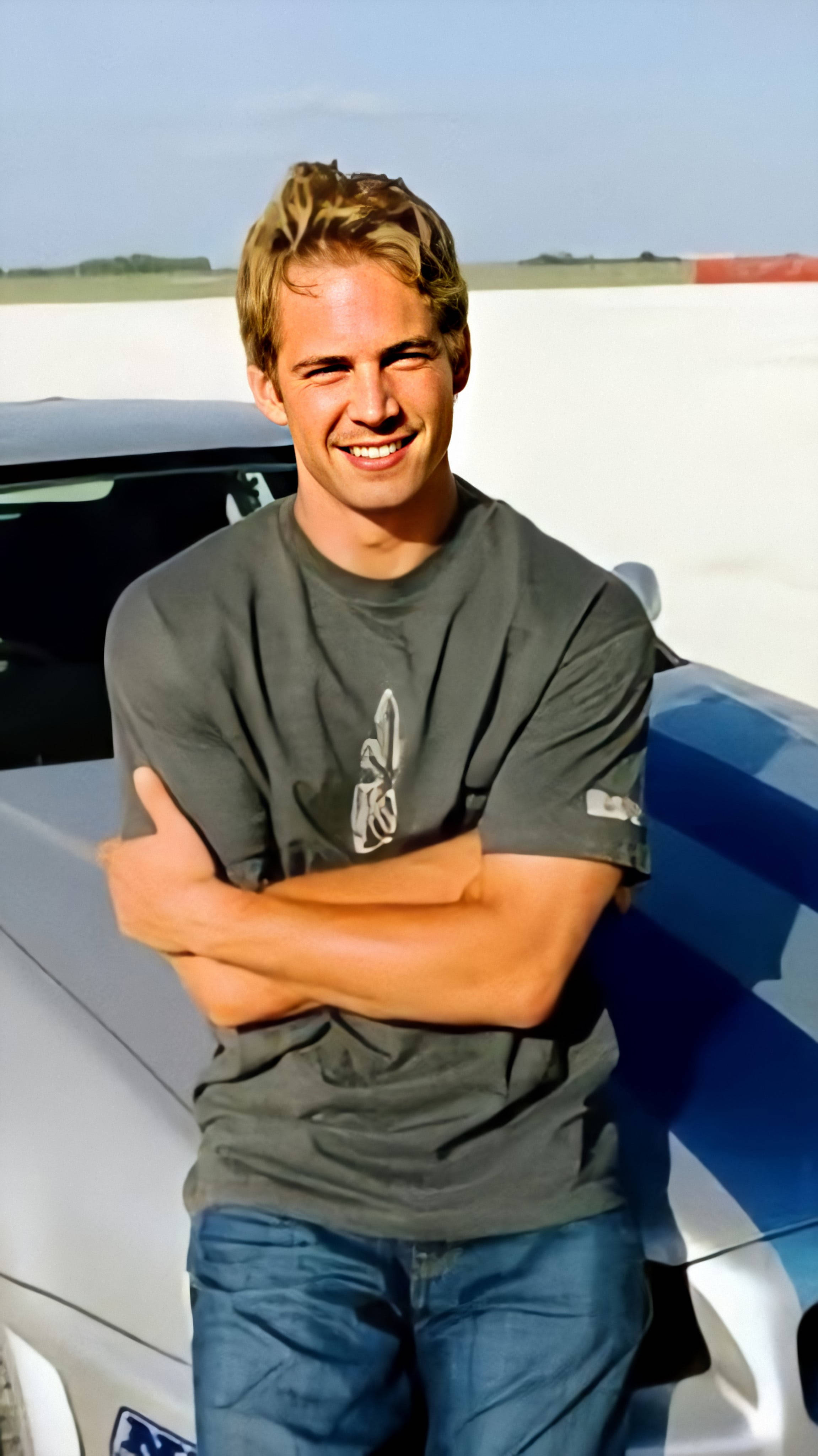 Paul Walker 桌布 壁紙 — 一個微笑的男人，穿著灰色T恤，在陽光明媚的賽道上自信地靠在一輛藍白賽車上。