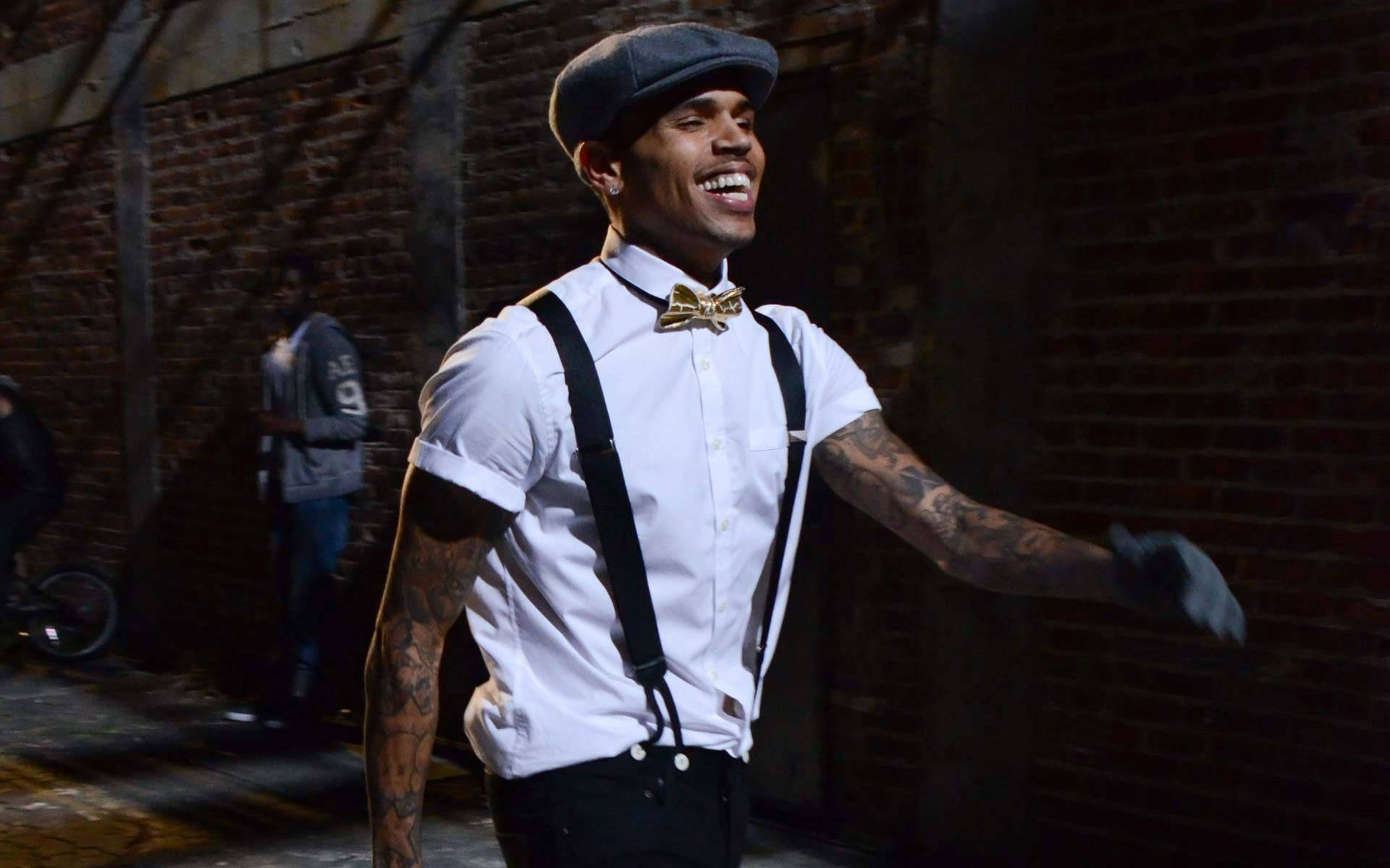 Chris Brown 桌布 壁紙 — 一個微笑的男人，穿著白襯衫、黑色吊帶和帽子，在夜晚對著磚牆充滿活力地跳舞。