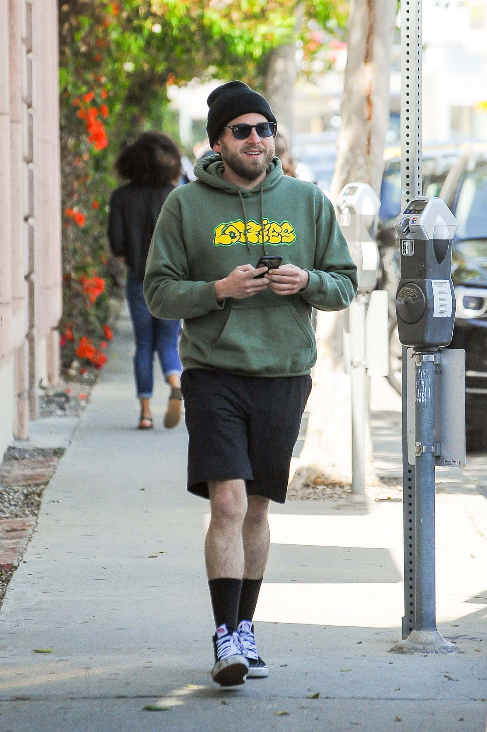 jonah hill 桌布 壁紙 — 一個微笑的男人，穿著綠色連帽衫和毛帽，在陽光明媚的人行道上邊走邊看手機。
