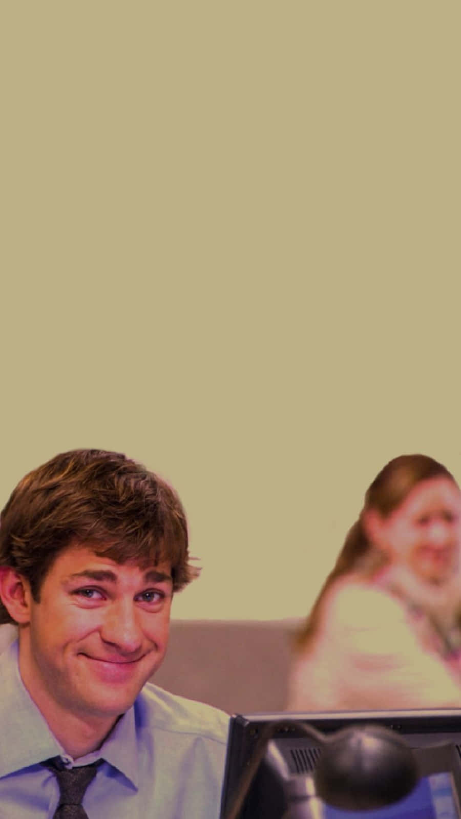 john krasinski 桌布 壁紙 — 一個微笑的男人，穿著襯衫打領帶，坐在電腦前面，後面有個模糊的女人，背景是純色米白牆。
