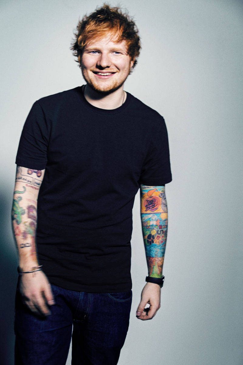 Ed Sheeran 桌布 壁紙 — 一個微笑的男人，紅髮，雙臂有彩色刺青，站在純灰色背景前，穿著黑色T恤和牛仔褲。