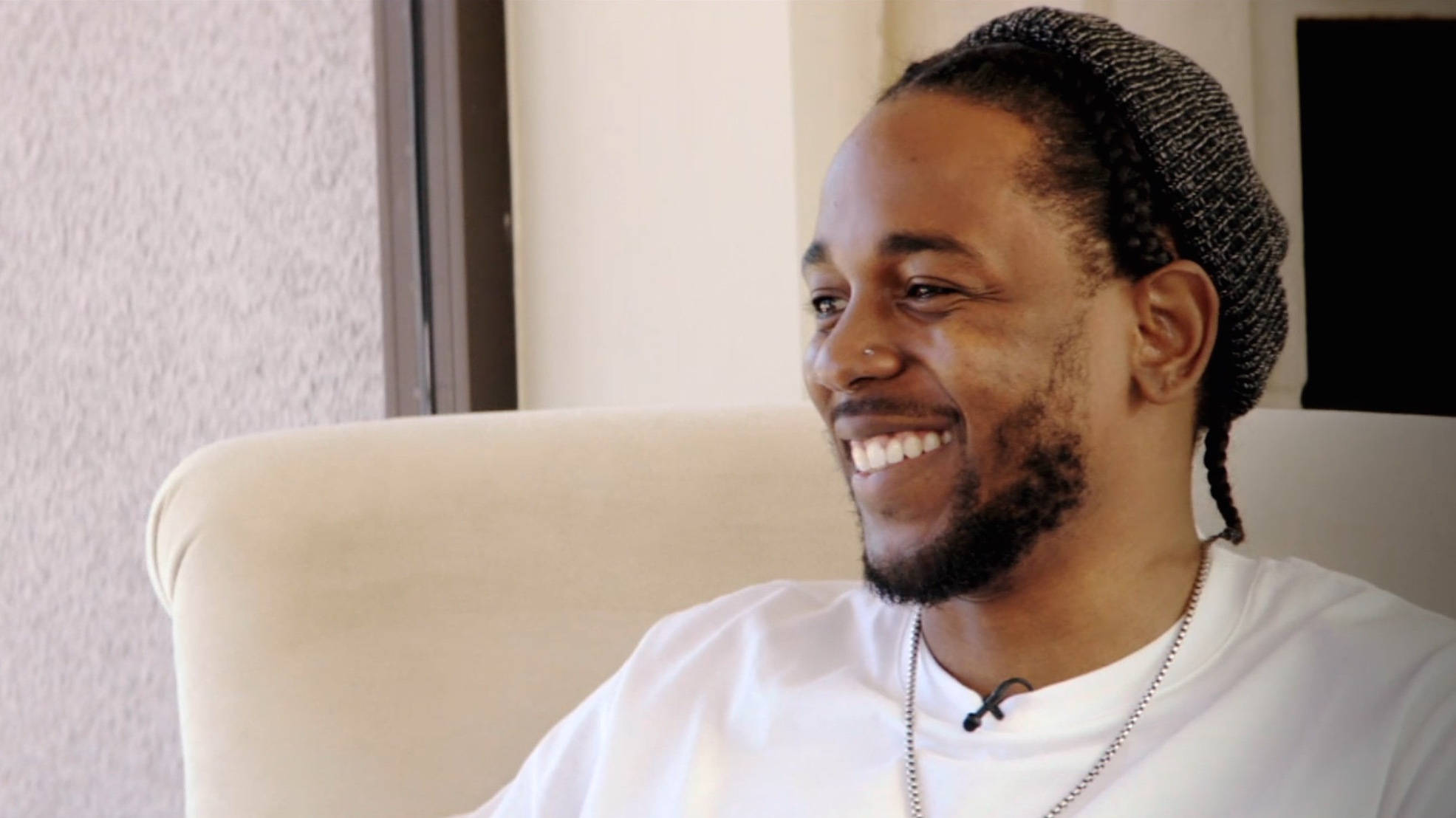 Kendrick 桌布 壁紙 — 一個微笑的男人，編著辮子，戴著無邊帽，舒適地坐在室內的米色扶手椅上。