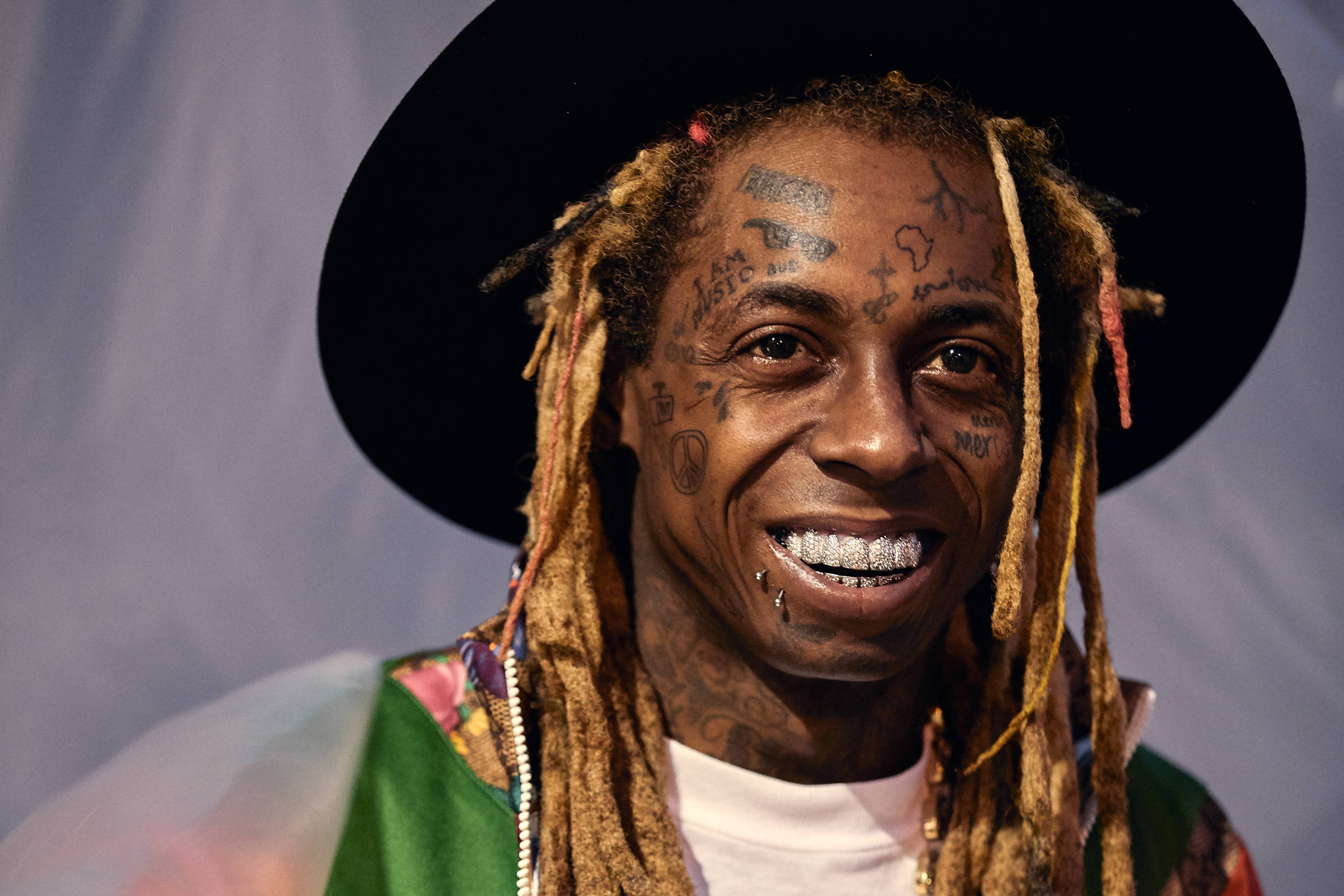 Lil Wayne 桌布 壁紙 — 一個微笑的男人，臉上紋身，留著辮子，戴著黑色帽子和彩色外套，背景樸素。