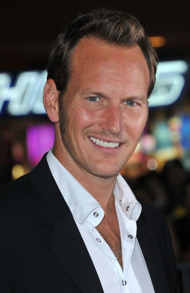 Patrick Wilson 桌布 壁紙 — 一個微笑的男人，藍眼睛，短黑髮，穿著黑色西裝外套，內搭白色開襟襯衫，背景是模糊的彩色。