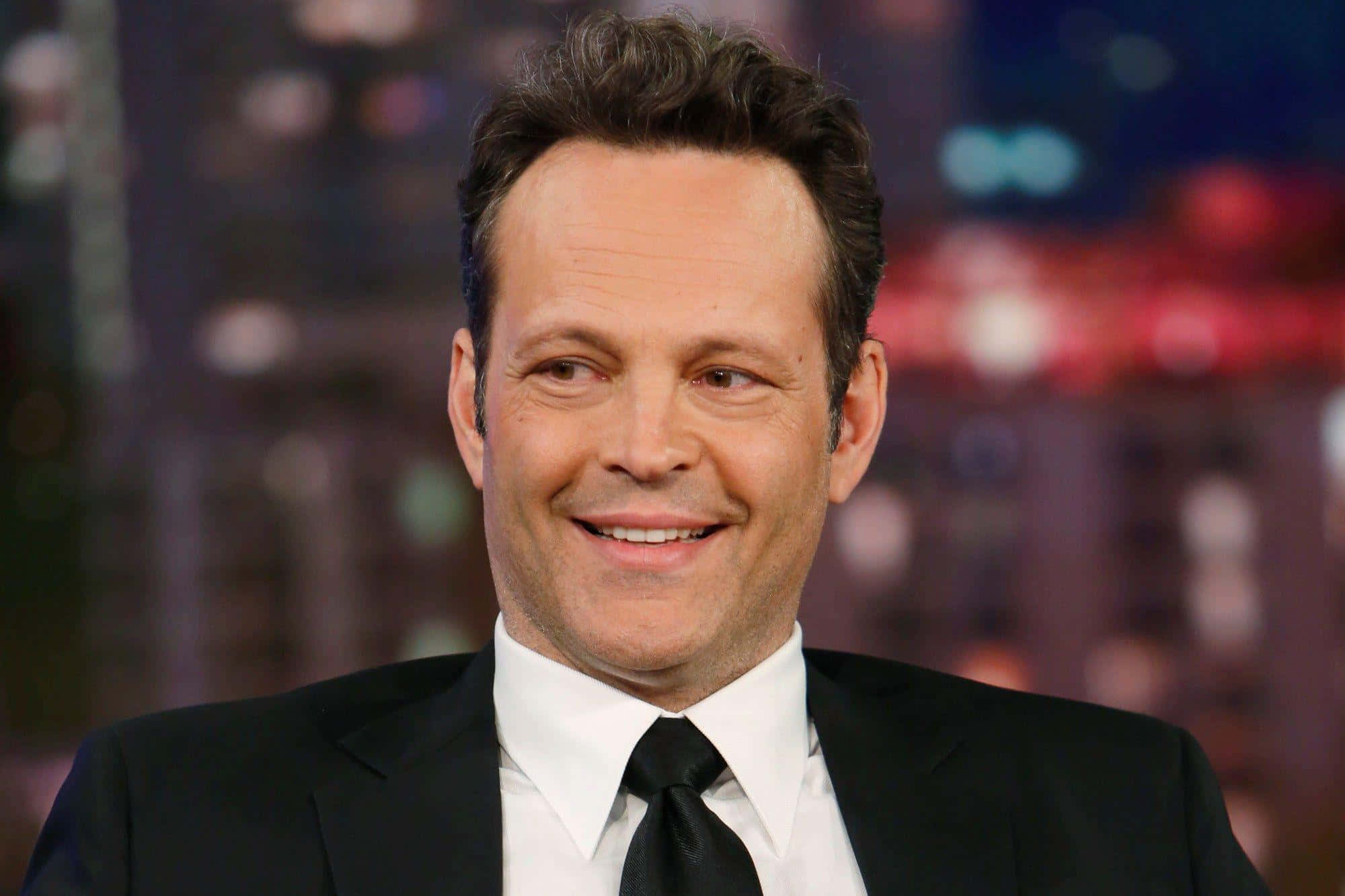 Vince Vaughn 桌布 壁紙 — 一個微笑的男人，身穿黑色西裝和領帶，背景是模糊的城市夜景，帶有色彩繽紛的散景燈光。
