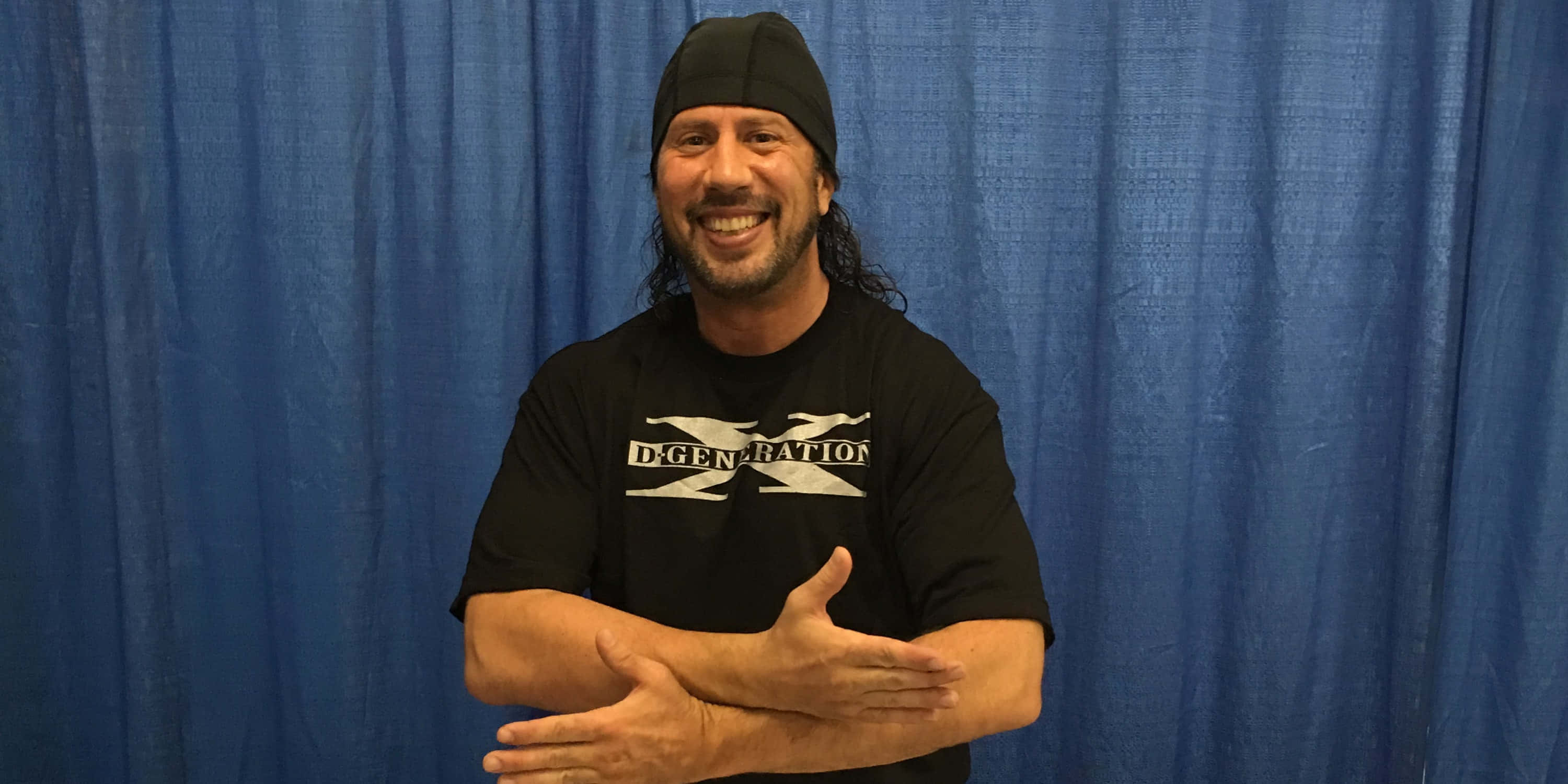 WWE Desktop 桌布 壁紙 — 一個微笑的男人，長長的黑髮和黑色無邊帽，自信地站在有紋理的藍色窗簾前，穿著帶有白色標誌的黑色T恤。