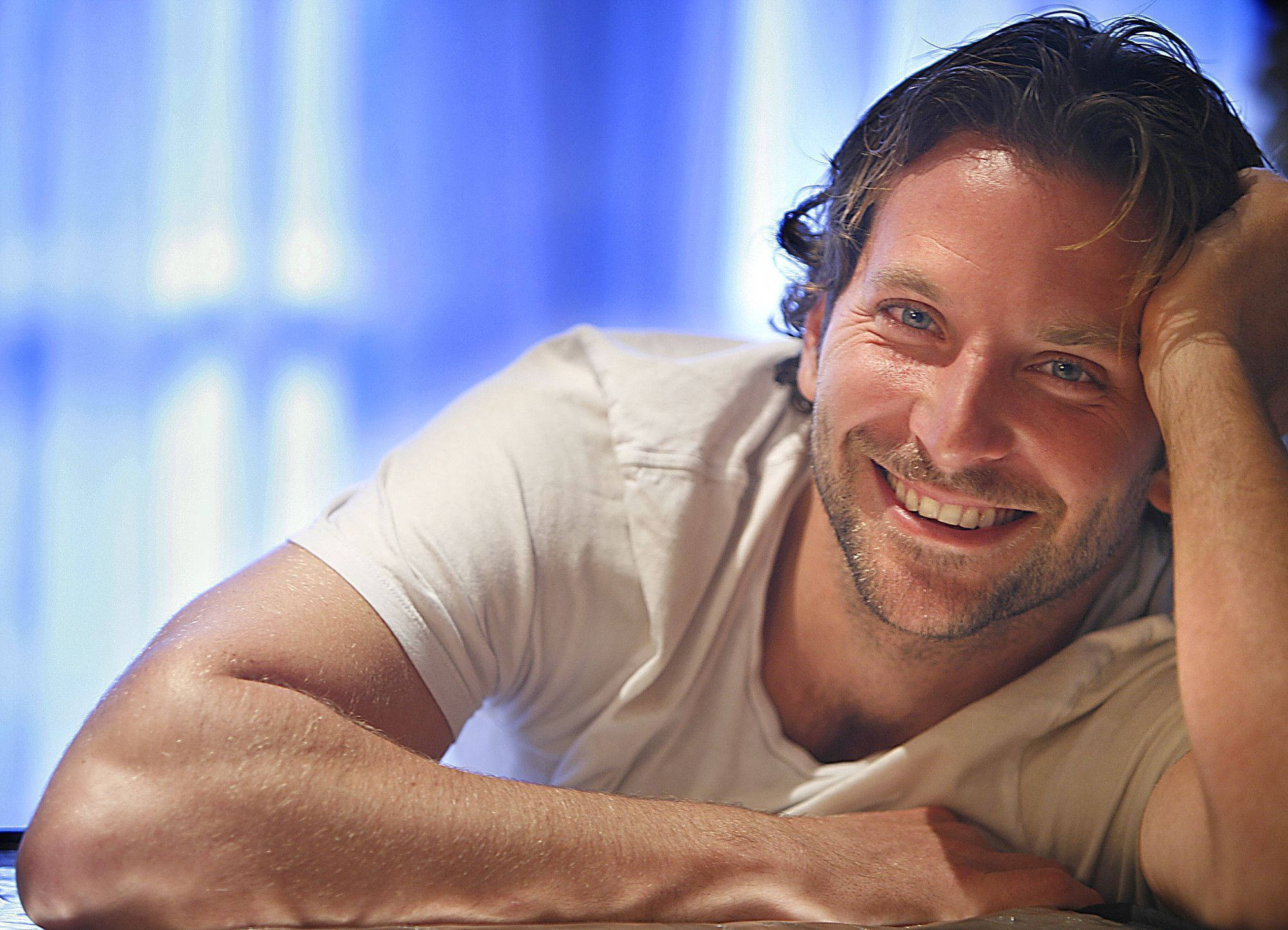 Bradley Cooper 桌布 壁紙 — 一個微笑的男人，頭髮蓬亂，將頭靠在手上，背景是柔和的藍色窗簾。