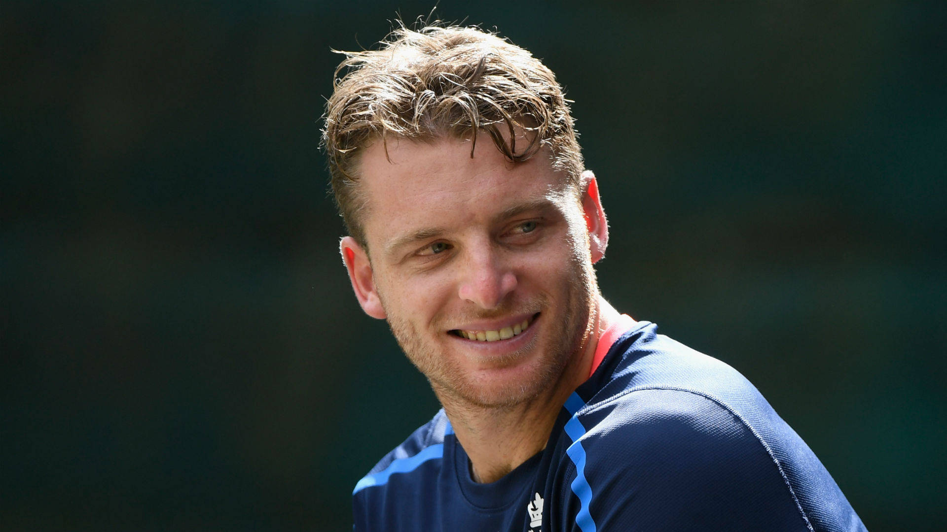 jos buttler 桌布 壁紙 — 一個微笑的男人，頭髮蓬亂，穿著藍色運動衫，在模糊的綠色背景下看向鏡頭外。