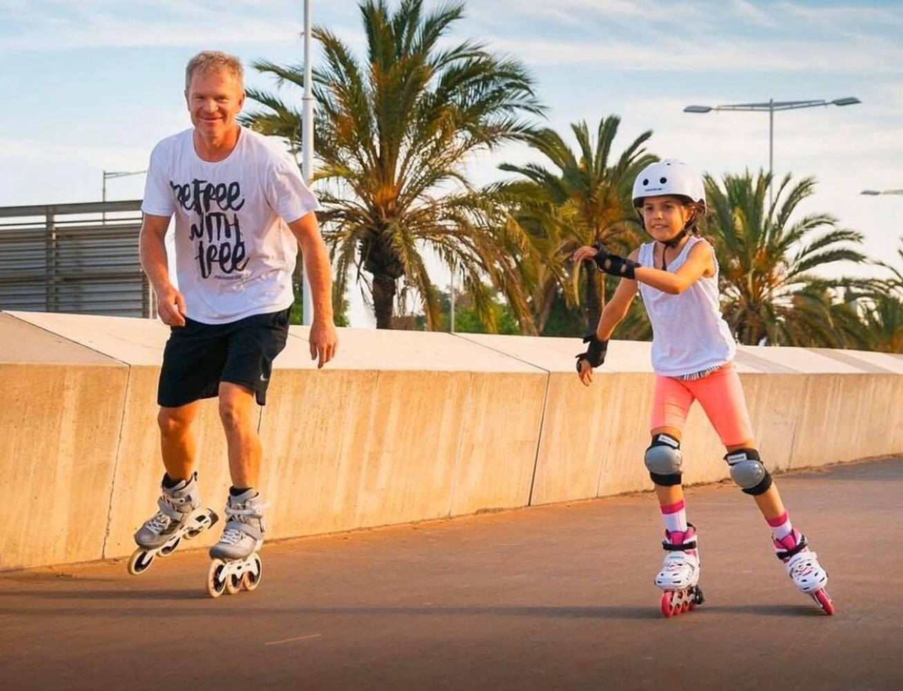 skating 桌布 壁紙 — 一個微笑的男人和一個年輕女孩在黃金時段，在棕櫚樹成蔭的鋪砌小徑上一起溜冰。