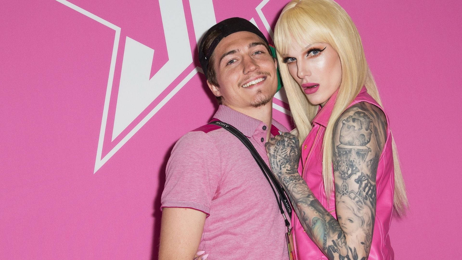 Jeffree Star 桌布 壁紙 — 一個微笑的男人和一個有刺青的金髮女人在一個帶有白色幾何標誌的亮粉紅色牆壁前緊密地擺姿勢。