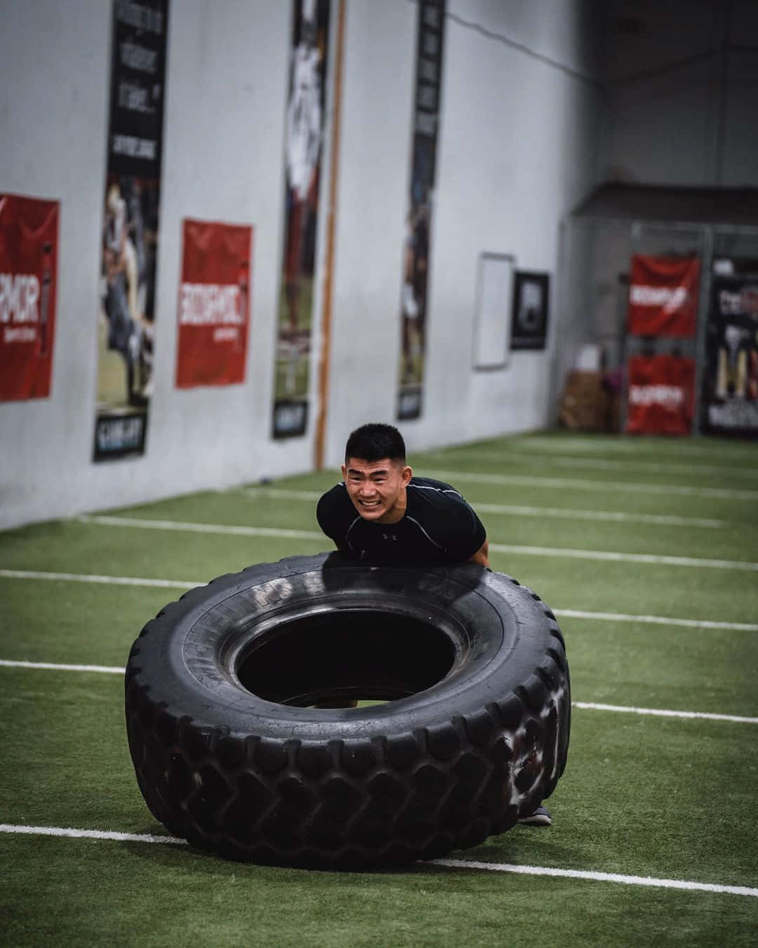 Workout 桌布 壁紙 — 一個微笑的男人在健身房內推著一個巨大的黑色輪胎穿過綠色草地場地，牆上掛著激勵海報。