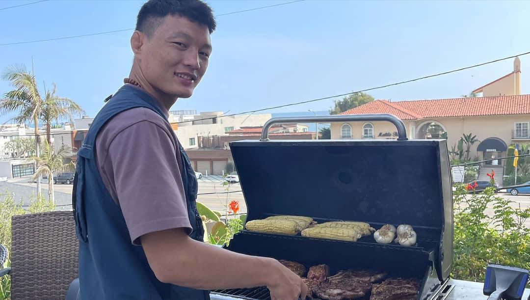 烹飪 桌布 壁紙 — 一個微笑的男人在陽光明媚的露臺上烤牛排和玉米，背景有棕櫚樹和地中海風格的房屋。