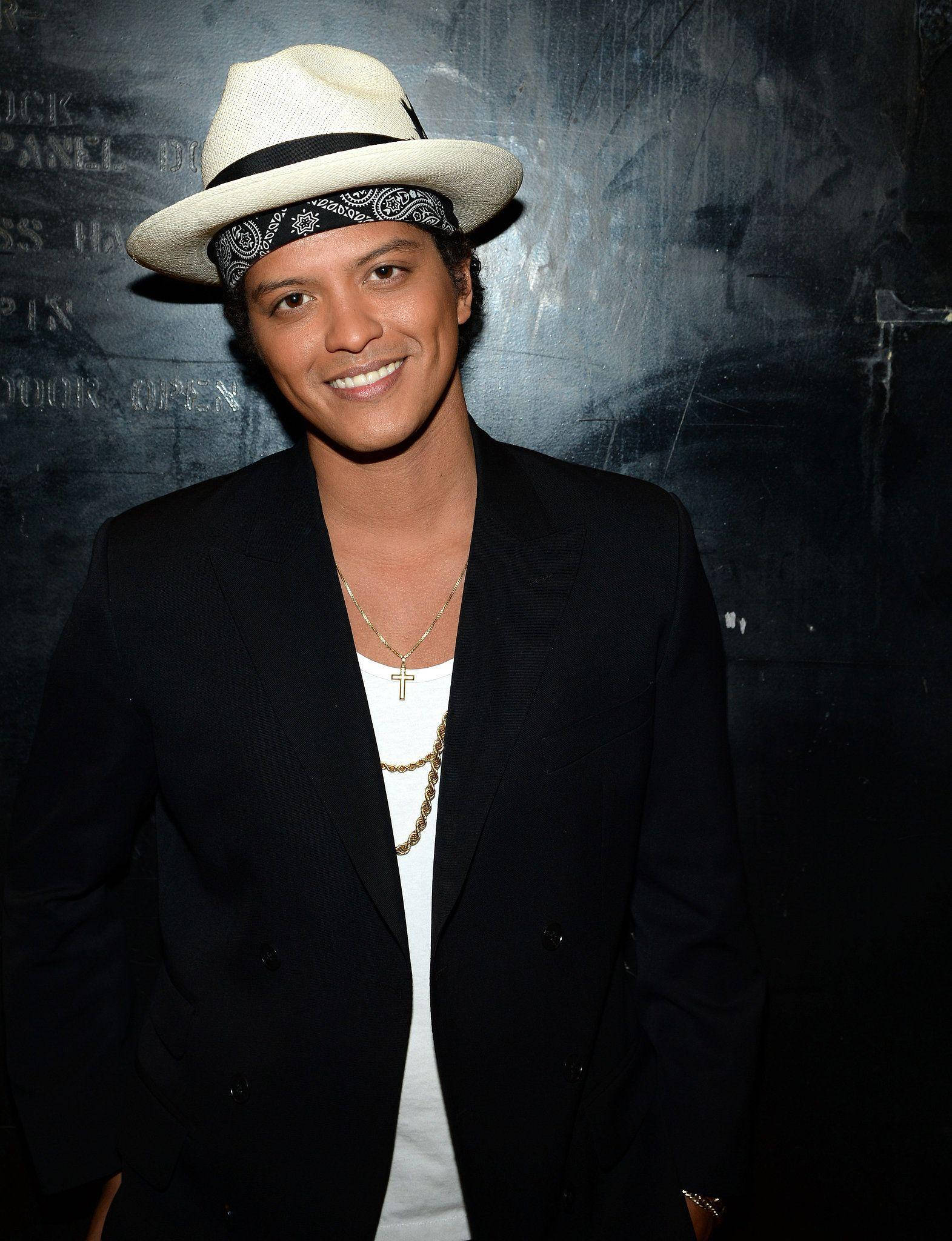 Bruno Mars 桌布 壁紙 — 一個微笑的男人戴著白色軟呢帽和頭巾，在帶有微弱塗鴉的深色紋理牆壁前擺姿勢。