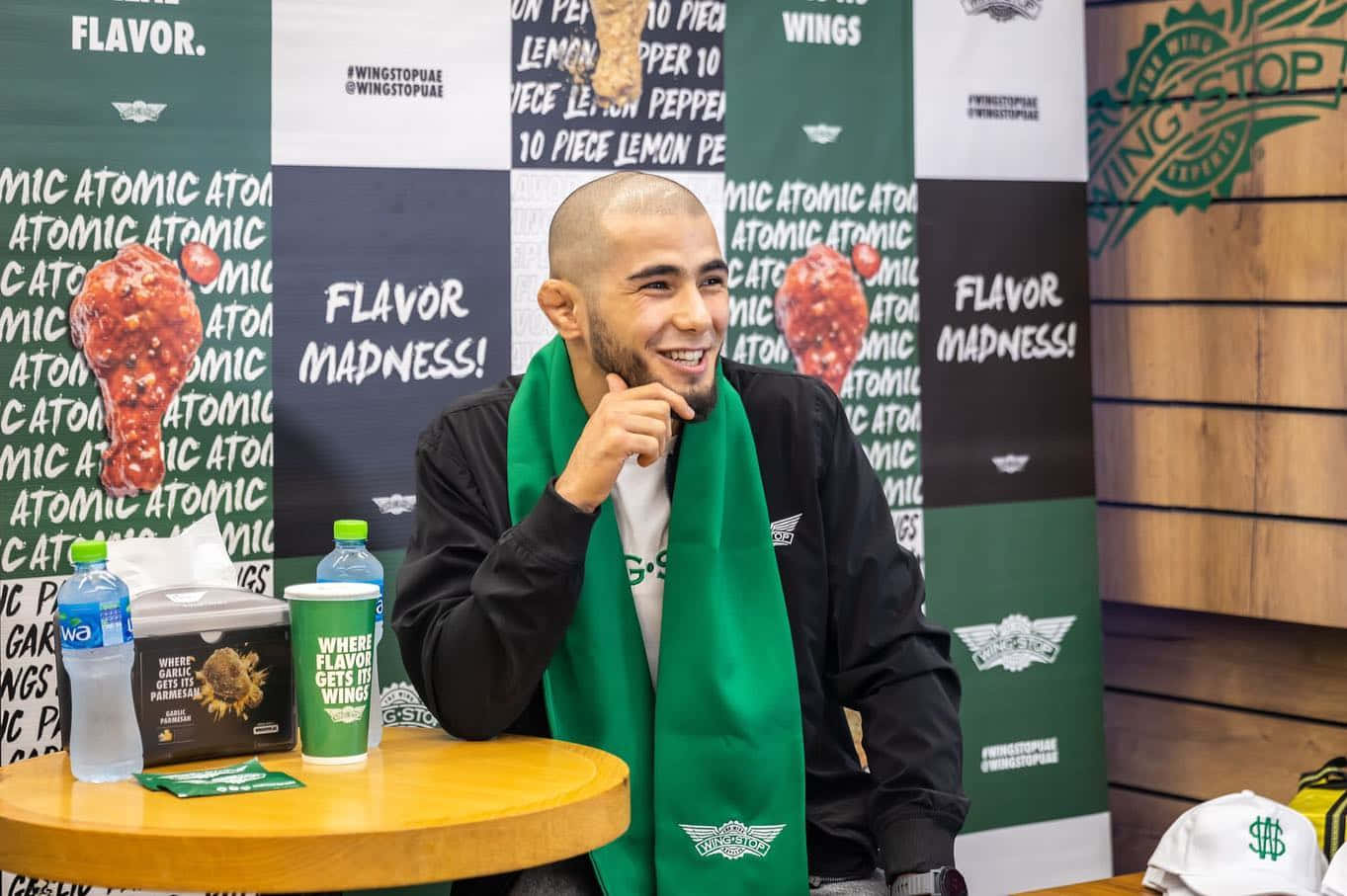 Celebrity 桌布 壁紙 — 一個微笑的男人戴著綠色圍巾，坐在一張後面有Wingstop品牌物品和食品包裝的桌子旁。