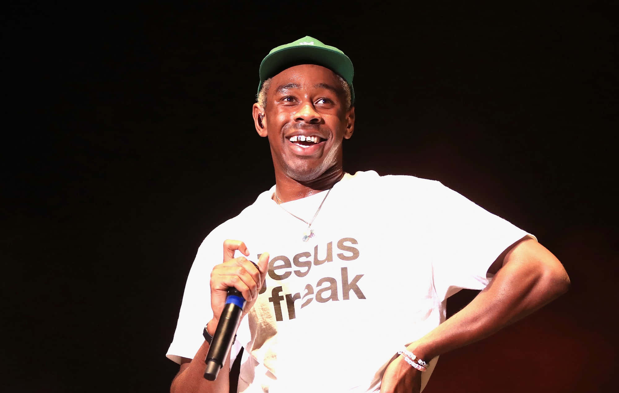 Rappers 桌布 壁紙 — 一個微笑的男人戴著綠色帽子和白色T恤，在黑暗的舞台上拿著麥克風。