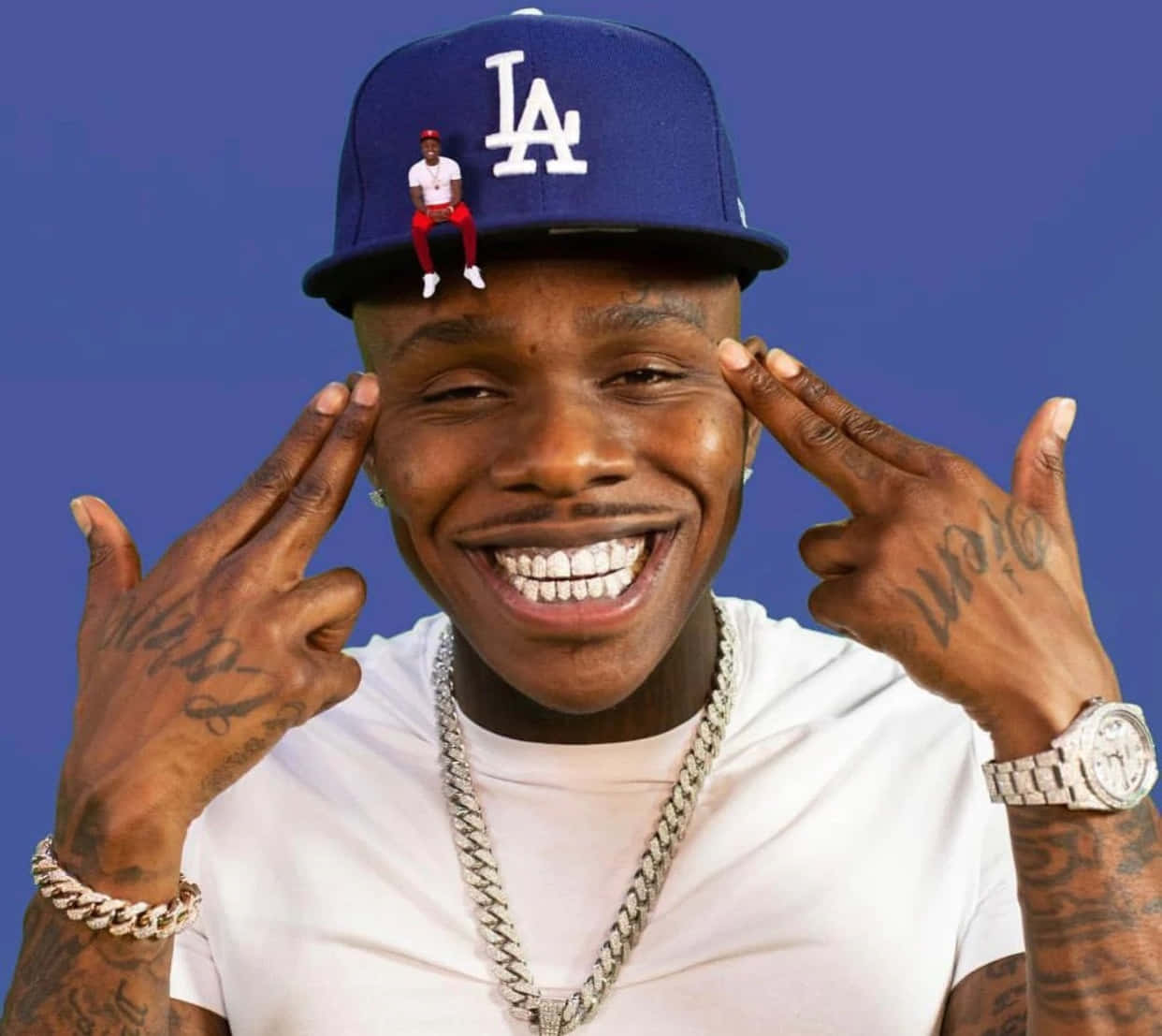 Dababy 桌布 壁紙 — 一個微笑的男人戴著藍色LA帽，上面坐著一個小人物，雙手框著他的臉，背景是純藍色。